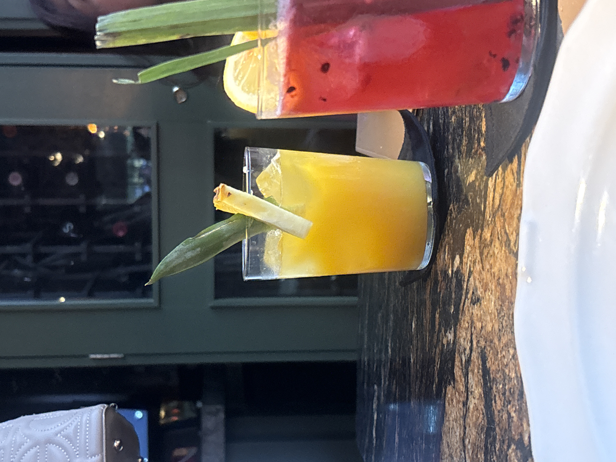 The image depicts two cocktails on a dark marble-patterned surface. The yellow cocktail is in a clear, square glass, garnished with a piece of fruit and a green leaf. The red cocktail is in a taller glass with a lemon slice and what appears to be a celery stalk. A partial view of a white object, possibly a plate or napkin, is on the right side of the frame. In the background, a window reflects the interior and exterior environment, with the dark colors. A purse is visible in the lower-left corner. The location seems to be in a restaurant or bar setting. The city of Puebla, Mexico is not discernible from the image.
