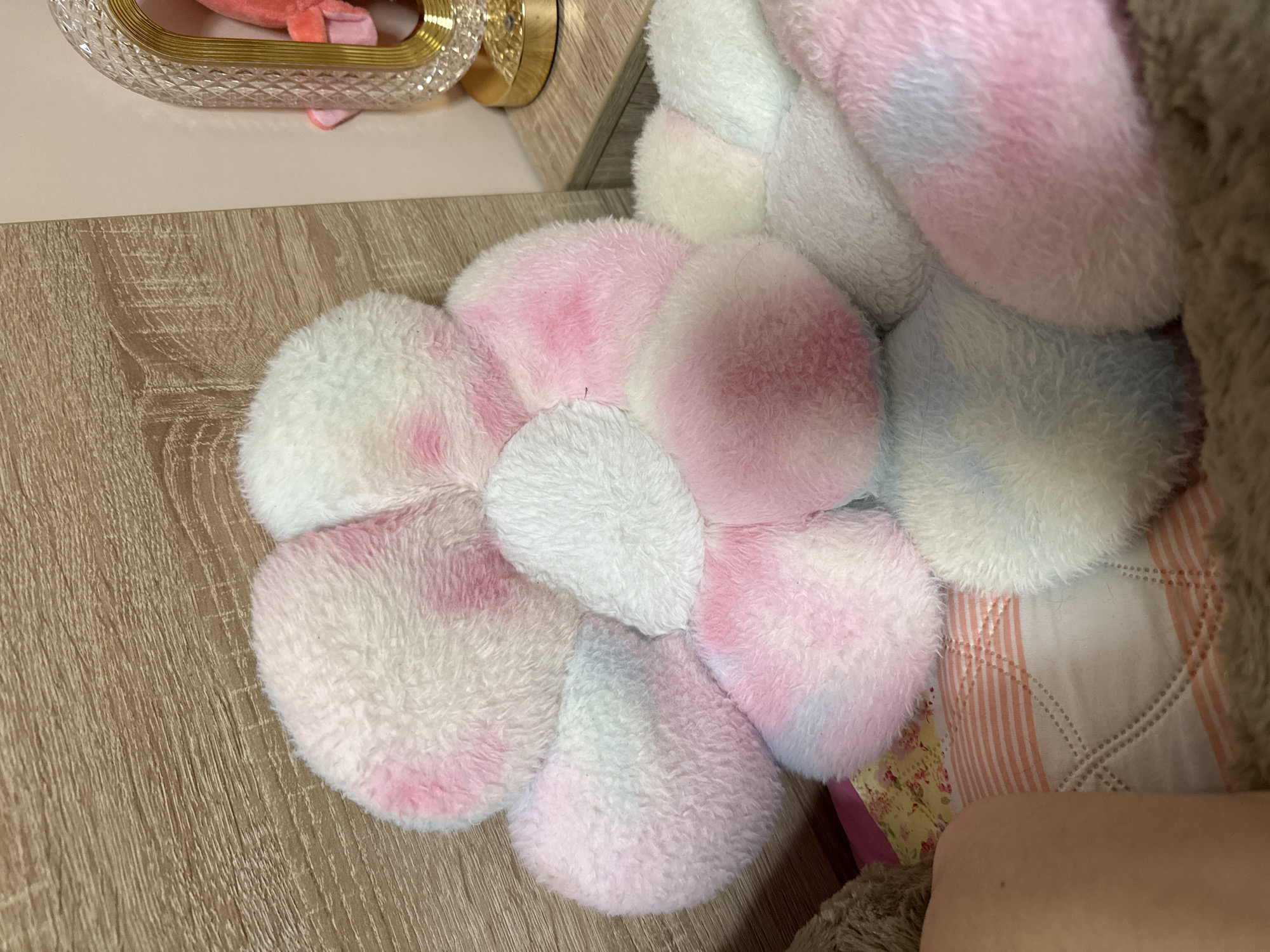 The image shows a fluffy flower-shaped cushion with pink and white petals. It is resting on a wooden surface. The cushion is partially obscured by a larger fluffy object on the right side. Part of a bed or bedding with a floral pattern is also visible near the edge of the cushion. To the left, a decorative glass and gold container sits atop a light-colored surface. There is no information to indicate it is taken in Iarebichna, Bulgaria.