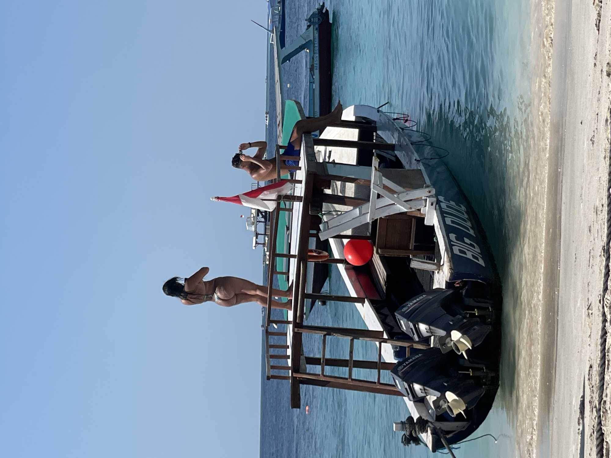 On a sunny day, a woman wearing a bikini stands on the top deck of a wooden and blue boat, looking away from the camera. To her right, a man in blue shorts sits on the same deck, holding his sunglasses and looking down. The boat, labeled "BIG DUDE" on its side, has two outboard motors at the stern and is anchored near a sandy beach. The water is a clear turquoise, and several other boats are visible in the distance. A red life preserver and a red ball are on the boat's deck. The scene suggests a recreational setting, likely a tropical beach destination.