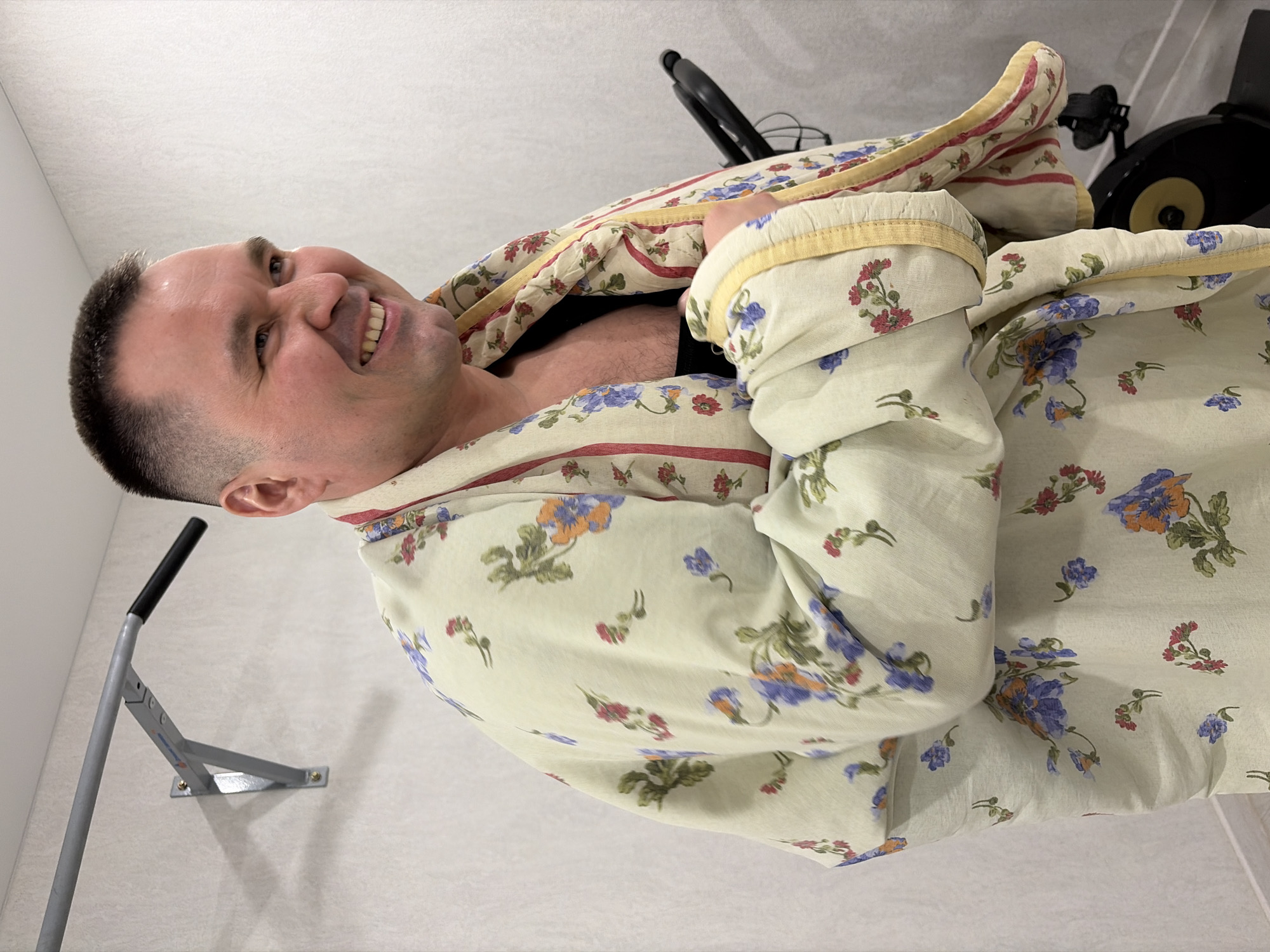 A man is lying in bed, covered with a floral-patterned blanket. He is smiling and appears to be in a relaxed environment. In the background, there is exercise equipment, including a pull-up bar mounted on the wall and an exercise bike. The setting appears to be a residential interior. There is no confirmed location or connection to the city of Sarapul, Russia.