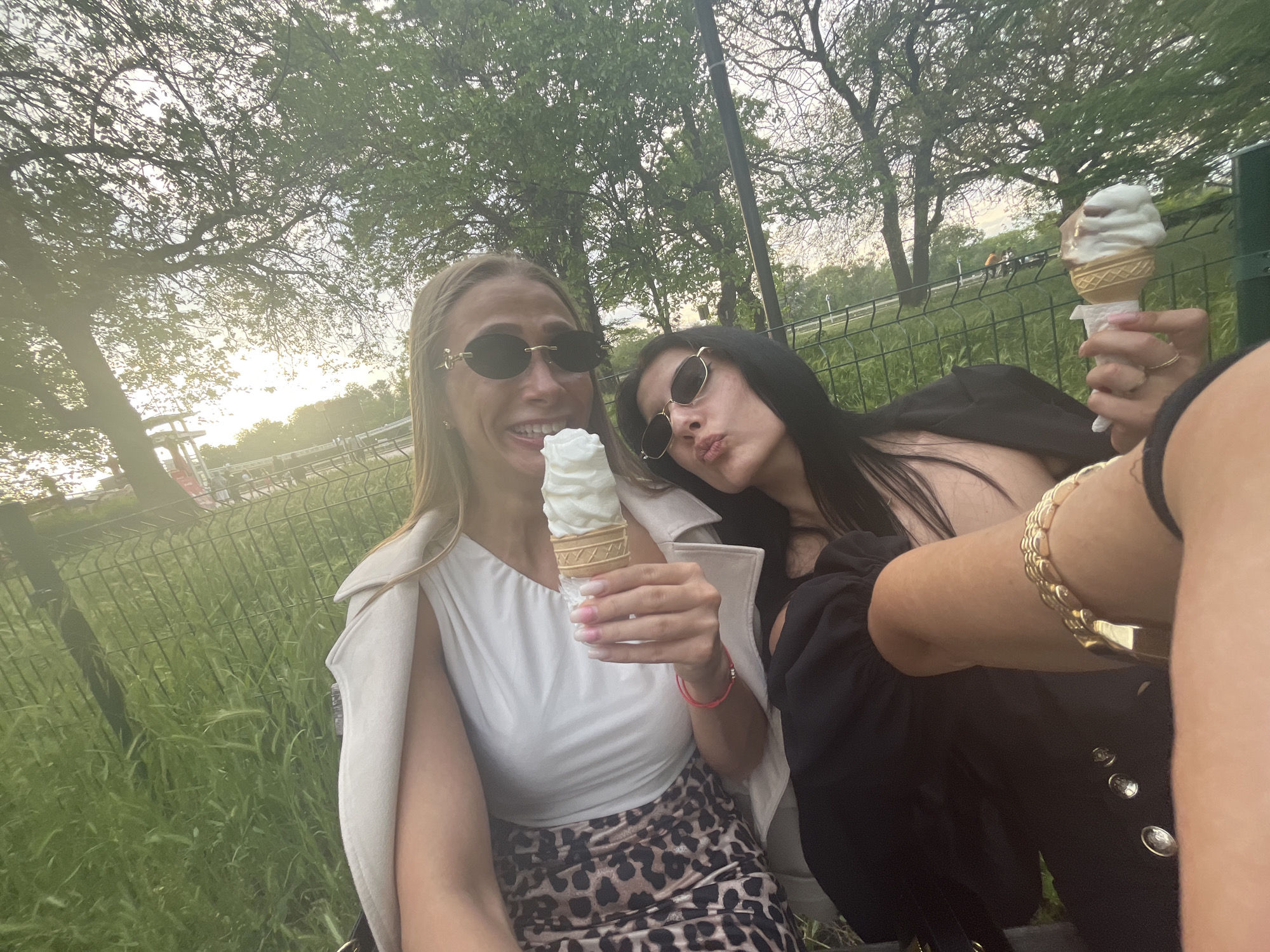 Two women are seated outdoors, possibly in a park, each holding an ice cream cone. The woman on the left has blonde hair, wears sunglasses, a white top, a beige jacket, and a leopard-print skirt. She is smiling and holding a white ice cream cone. The woman on the right has dark hair, wears sunglasses, and a black outfit. She is posing with puckered lips while holding a cone with both vanilla and chocolate ice cream. The background features a green grassy area with a metal fence and trees. The lighting suggests it is either early morning or late afternoon. The location cannot be verified as being in Plovdiv, Bulgaria.
