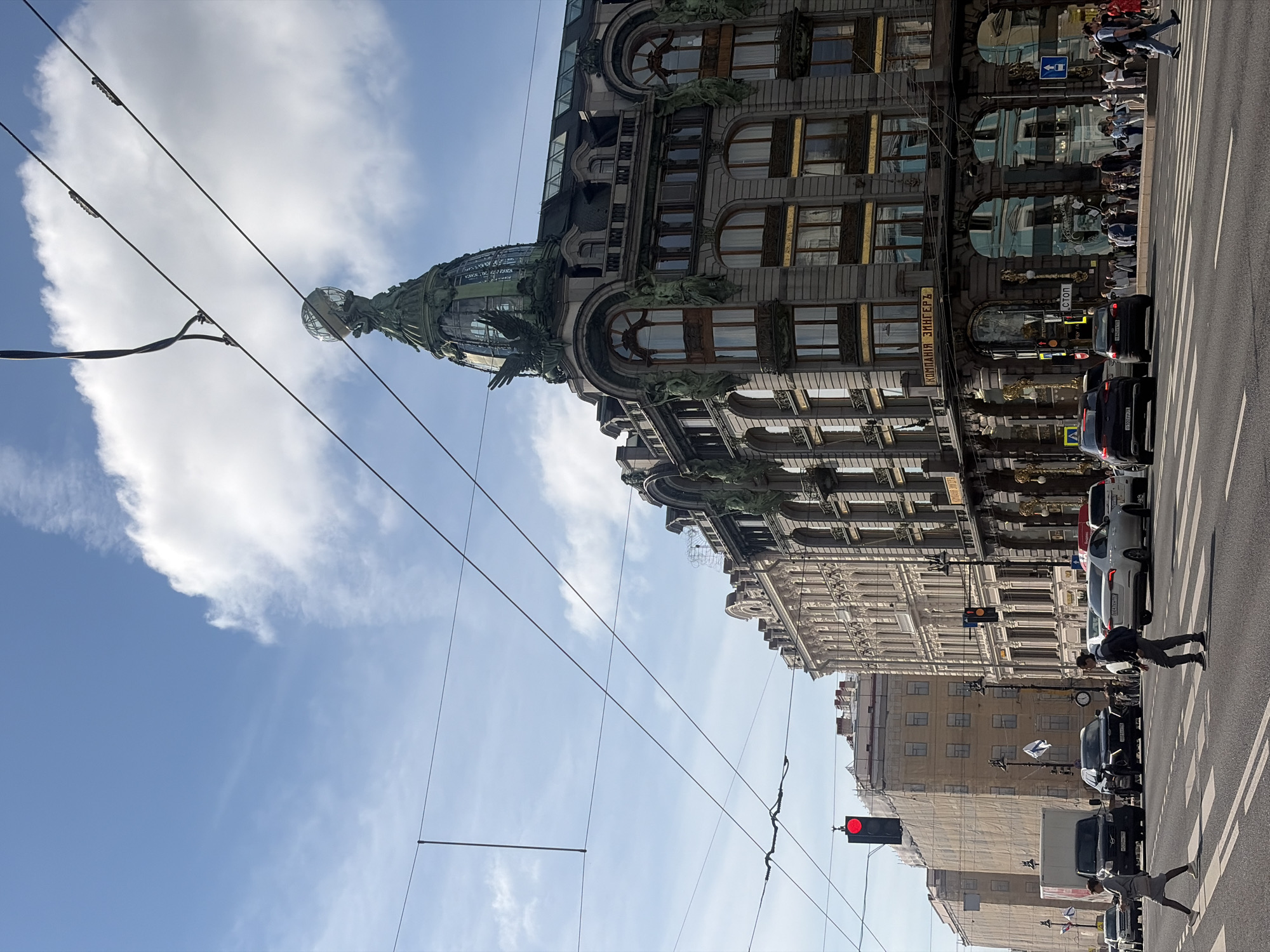 This is a street-level, eye-level, slightly angled, medium shot of Nevsky Prospect in Saint Petersburg, Russia, on a sunny day with scattered clouds. The main subject is the Singer House (Dom Zinger) building, a prominent Art Nouveau architectural landmark with a distinctive glass dome topped by an eagle sculpture. The building's facade features large arched windows with golden detailing and decorative sculptures.
In the foreground, a street intersection with traffic lights, a pedestrian crossing, and several vehicles, including cars and a white van, are visible. Pedestrians are crossing the street, some walking briskly. The sky above is blue with some white clouds. Overhead, numerous electrical wires crisscross the frame, suggesting the presence of trolleybus or tram lines. To the left of the Singer House, another building with a lighter facade is partially visible, and further left, a building is covered in scaffolding and protective sheeting. The overall scene depicts a busy urban environment with a focus on the historical architecture and daily life of Saint Petersburg.