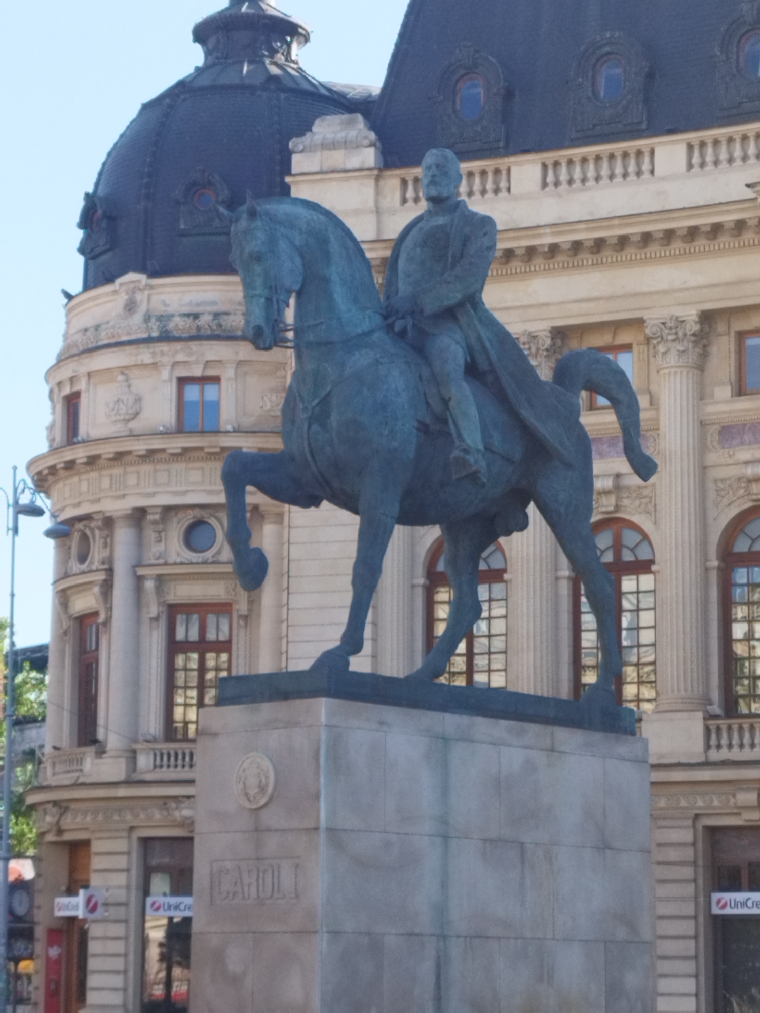 The image depicts the equestrian statue of King Carol I, a prominent landmark located in Bucharest, Romania, specifically in front of the National Museum of Art of Romania. The statue, rendered in a dark, weathered material, shows the king in military attire, seated atop a rearing horse. The horse is captured in mid-gallop, its front legs elevated. The base of the statue is a large, stone pedestal. The inscription "CAROLI" is visible on the front of the pedestal, a clear indication of the statue's subject.
Behind the statue, the facade of a grand, classical building is visible. This building features a dark, domed roof section on the left, and a lighter colored, multi-story structure with numerous windows, columns, and ornate architectural details. The windows are tall and rectangular, with some arched tops. The building's architecture suggests it is a significant public or historical edifice. Sunlight illuminates the scene, casting some shadows and highlighting the details of the statue and the building.
On the left side of the image, a street lamp with multiple lights is partially visible. There are also indications of other elements of the urban environment, such as what appears to be part of another building or structure on the far left, and signage with the "UniCredit" logo on the building facade below the statue's pedestal. The sky is a clear, light blue, suggesting a sunny day. The overall impression is one of historical grandeur and public space.