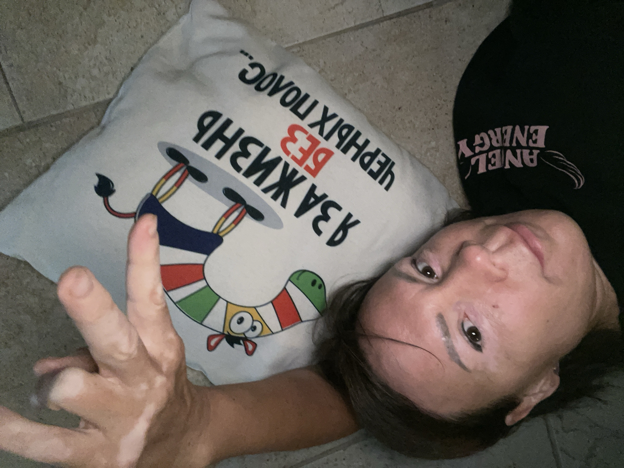 A woman lies on her left side, facing a light-colored pillow with a graphic and text. She is wearing a black t-shirt with pink text and graphics on the left side of her chest. Her right arm is extended and resting on the pillow, with her hand visible. The pillow features a cartoon drawing of an animal with a striped body, large eyes, and red ears, standing on black circles. The text on the pillow, written in Russian, translates to "I wish for a life without black stripes." The woman's face is partially visible in the upper left quadrant of the image. The environment appears to be indoors, with a tiled floor visible in the background. The image is a selfie taken from a high angle, looking down at the woman and the pillow. This scene is not in the city of Sochi, Russia.