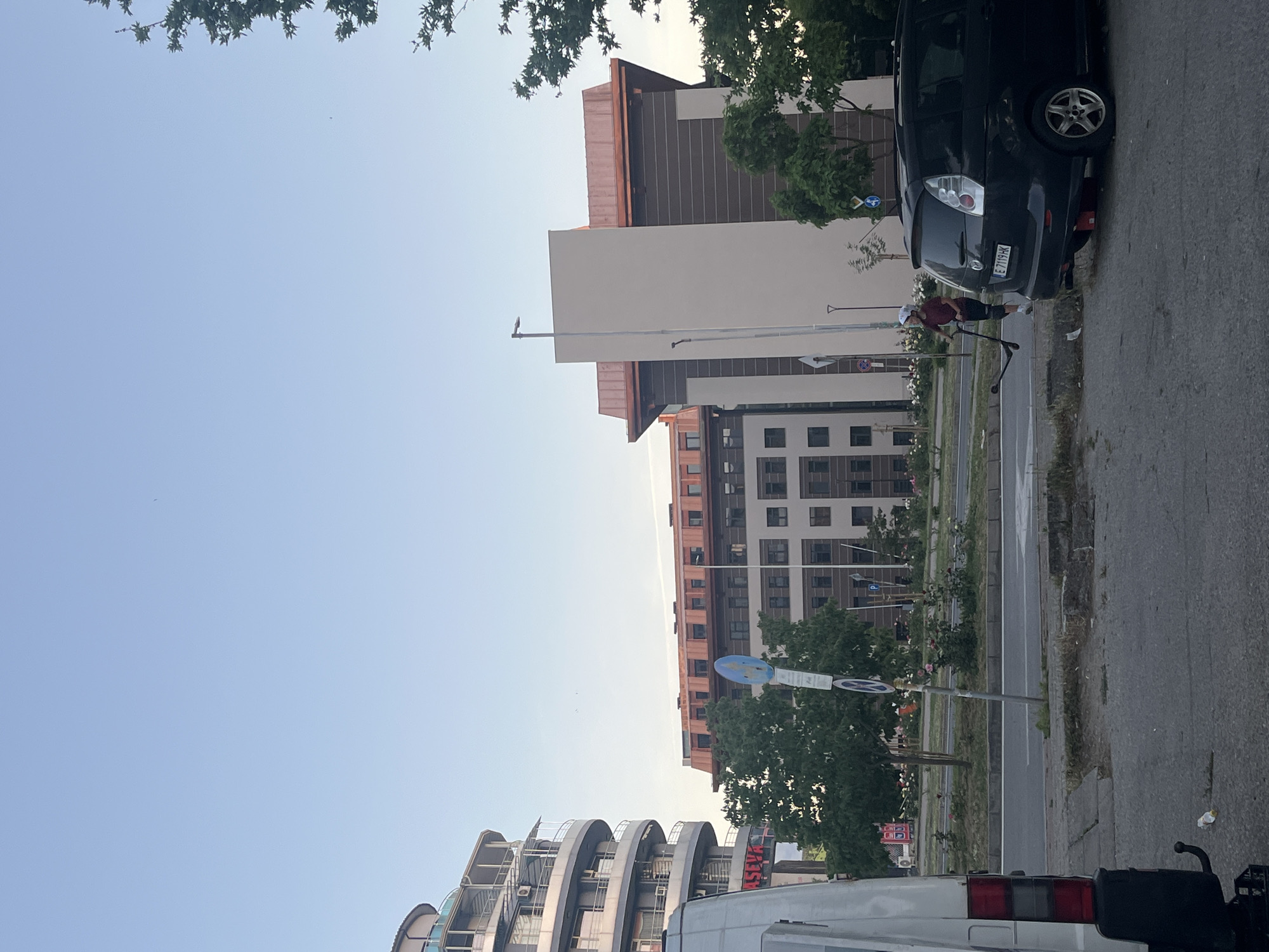 The image presents an urban street scene in Blagoevgrad, Bulgaria. Several buildings are visible, including a modern apartment complex with curved balconies and a larger building with a copper-colored roof. A dark car is parked along the street, its rear partially visible. A person on a scooter is adjacent to the car. A white van occupies the lower portion of the frame, facing away. Street signs, including parking signs, are visible. The street is paved and contains a sidewalk. The sky is light blue. Trees are scattered throughout the scene.