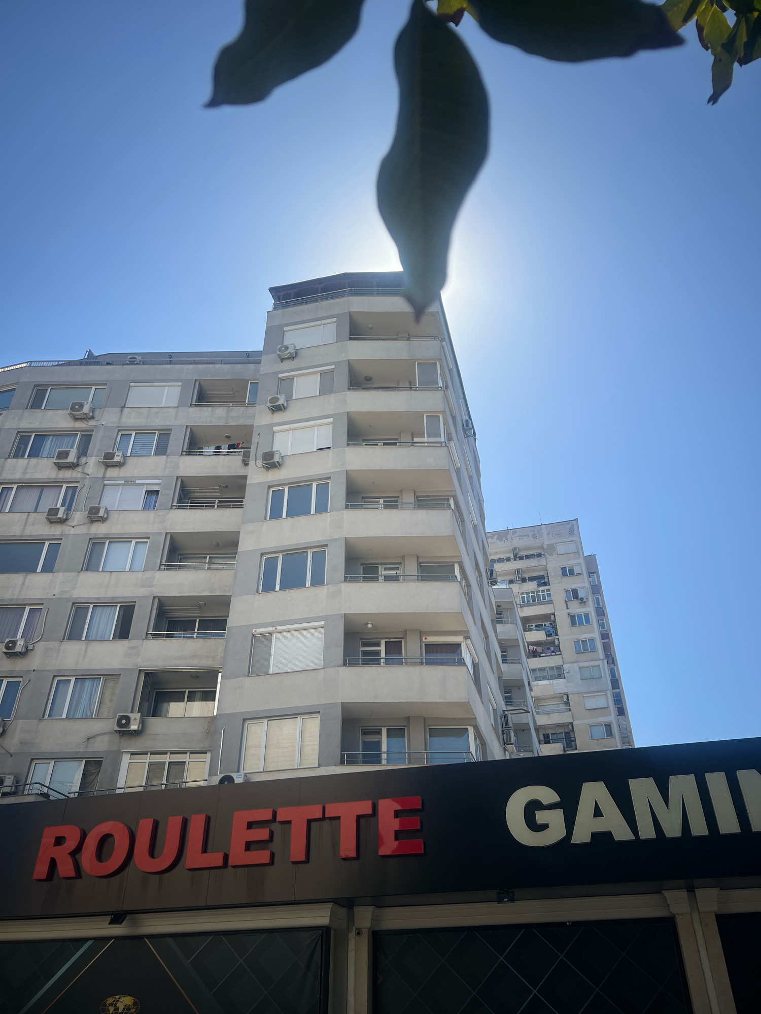 The image shows a close-up of a dark sign with the words "ROULETTE GAMING" in large red and white letters, respectively. Above the sign, a tall, grey apartment building with multiple balconies and windows is visible. The sun is shining brightly, creating a lens flare effect and casting shadows. In the foreground, out of focus, are parts of leaves from a tree or plant. The scene is likely in an urban environment, and the presence of the "ROULETTE GAMING" sign suggests it is a commercial establishment. Based on the architectural style and the overall appearance, the location appears to be in Plovdiv, Bulgaria, specifically along the main pedestrian street, Knyaz Alexander I Street, near the central square.