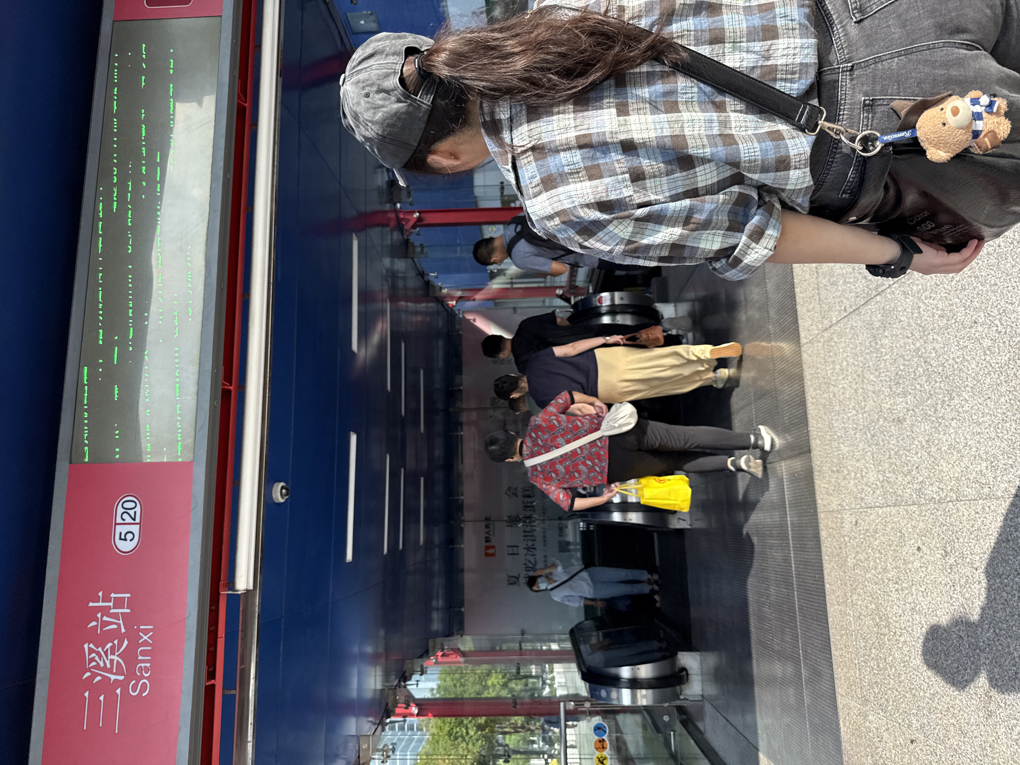 The image shows the entrance to the Sanxi metro station in Tianhe District, China. A pink sign above the entrance reads "三溪站 Sanxi 5 20". Below the sign, a digital display shows green text and symbols. Several people are walking into the station, some are going up an escalator, while others are walking down. One person in the foreground, seen from the back, is wearing a plaid shirt and a baseball cap. The scene is well-lit, likely during the day, with sunlight casting shadows on the ground.