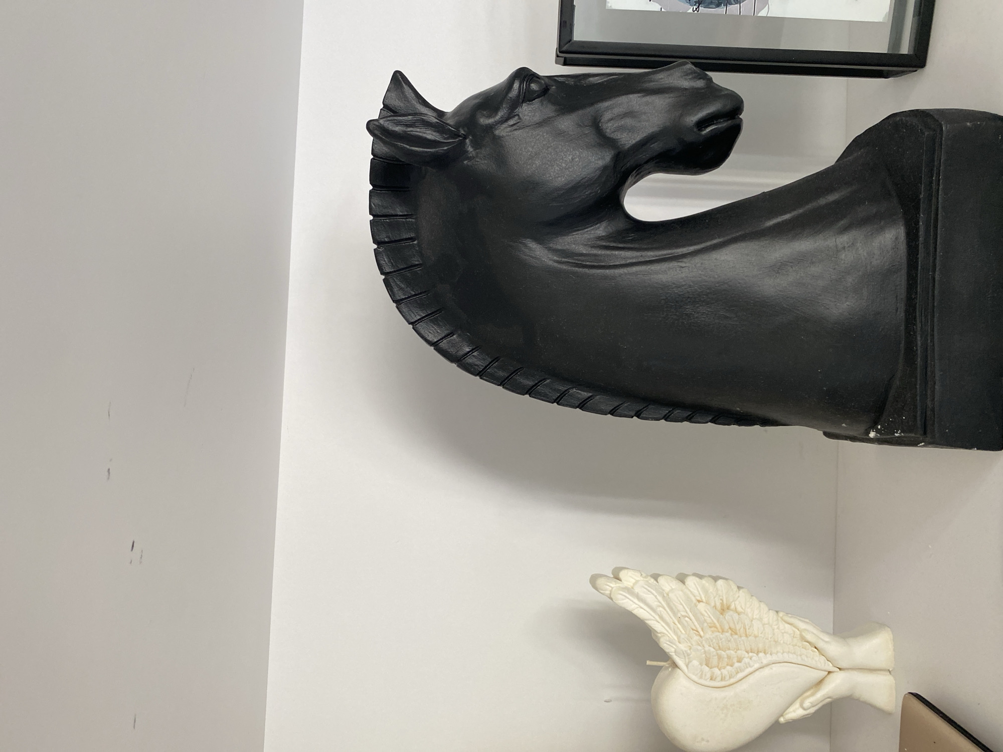 The image shows a white shelf against a white wall in a room. On the shelf, to the left, is a cream-colored candle shaped like a heart with angel wings and hands at its base. To the right of the candle is a large, black, matte sculpture of a horse's head, likely a chess piece, with a detailed mane and face. Further to the right, a portion of a framed picture is visible. The shelf itself appears to be made of a laminate material. The environment is clean and minimalist. There are a few faint dark marks on the wall above the shelf. The image does not provide any information about the city of Buk, Poland, or any specific location within it.