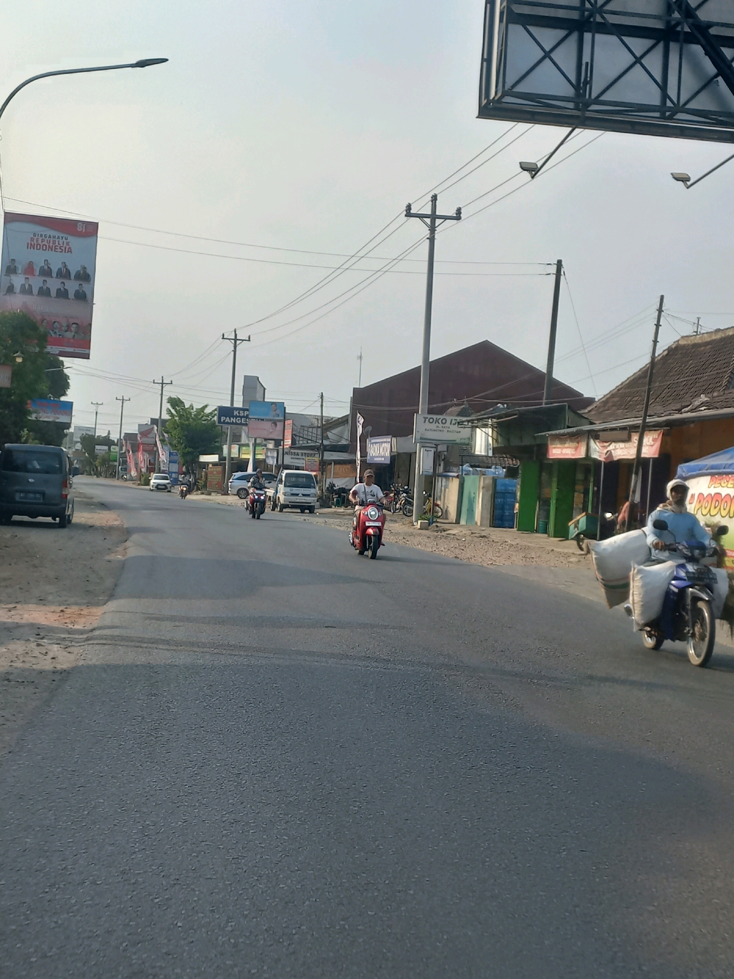 The image depicts a street scene in Baturetno, Indonesia, likely during daytime given the bright sky and sunlight. The road appears to be asphalted and shows signs of wear and tear. On the left side of the road, a dark-colored van is parked. Further down the road, several motorcycles and a white car are visible.
There are multiple commercial buildings lining the right side of the street. One building has a sign that reads "TOKO IDEAL" and "BATURETNO RAJATAN". Other buildings have various signs, some of which are difficult to read due to resolution or distance. There are also utility poles and power lines crisscrossing the sky.
In the foreground and mid-ground, several people are riding motorcycles. One person on a red scooter is in the left lane, while another individual on a blue motorcycle is in the right lane, carrying a large load wrapped in what appears to be sackcloth. Another motorcycle is seen further ahead in the left lane. A billboard is visible in the upper left corner of the image, featuring images of people and text. The overall impression is that of a busy local street with commercial activity and road traffic.