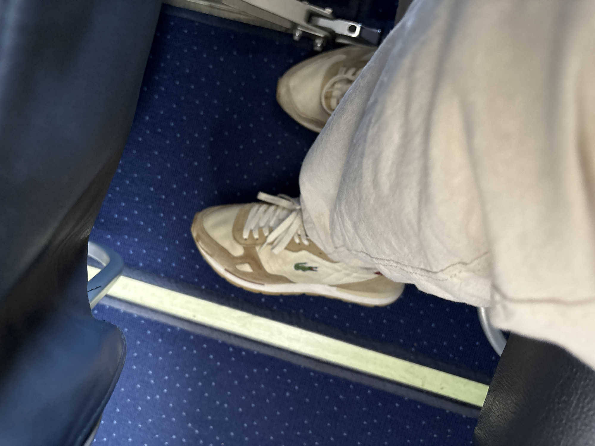 The image shows a person sitting on an airplane. The person is wearing white pants and Lacoste sneakers. The airplane's carpet is blue with small white dots. The seats are blue leather. The floor is narrow and the person's feet are close together.