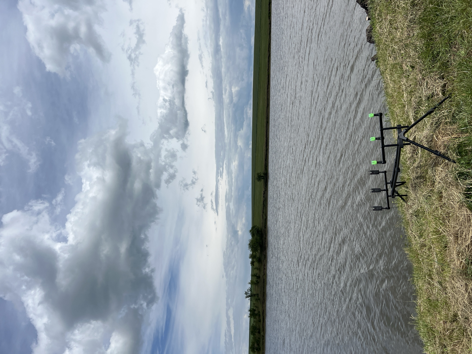 The image depicts a body of water, likely a lake or river, with a shoreline in the foreground. On the grassy bank, a black fishing rod pod with three green rod rests is set up. The water has ripples and appears calm. Beyond the water, a treeline with scattered bushes separates the water from a vast, gently sloping green field. The sky above is filled with dramatic, grey and white clouds, suggesting an overcast day. The scene is located in the village of Borychivka, Ukraine.