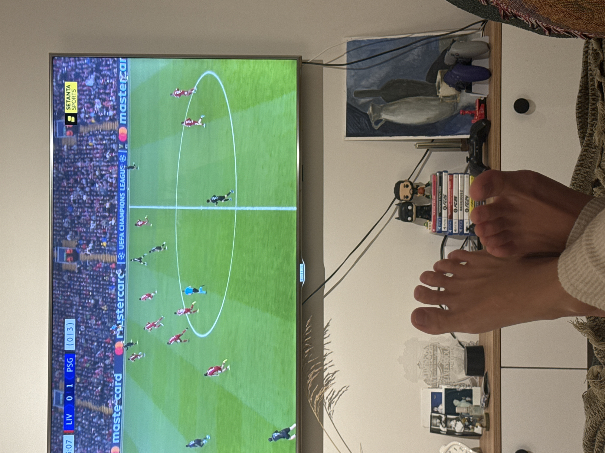 The scene captures a television displaying a soccer match between Liverpool and Paris Saint-Germain, with the score at 0-1 in favor of PSG. The game is being broadcasted by Setanta Sports. The TV is positioned above a white cabinet or shelf, which holds various items including video game cases, figurines, gaming controllers, a painting, a lamp, and photographs. A pair of human feet rests on the edge of the cabinet near the TV. The environment is indoors and appears to be a residential setting. The specific city and location remain unknown.