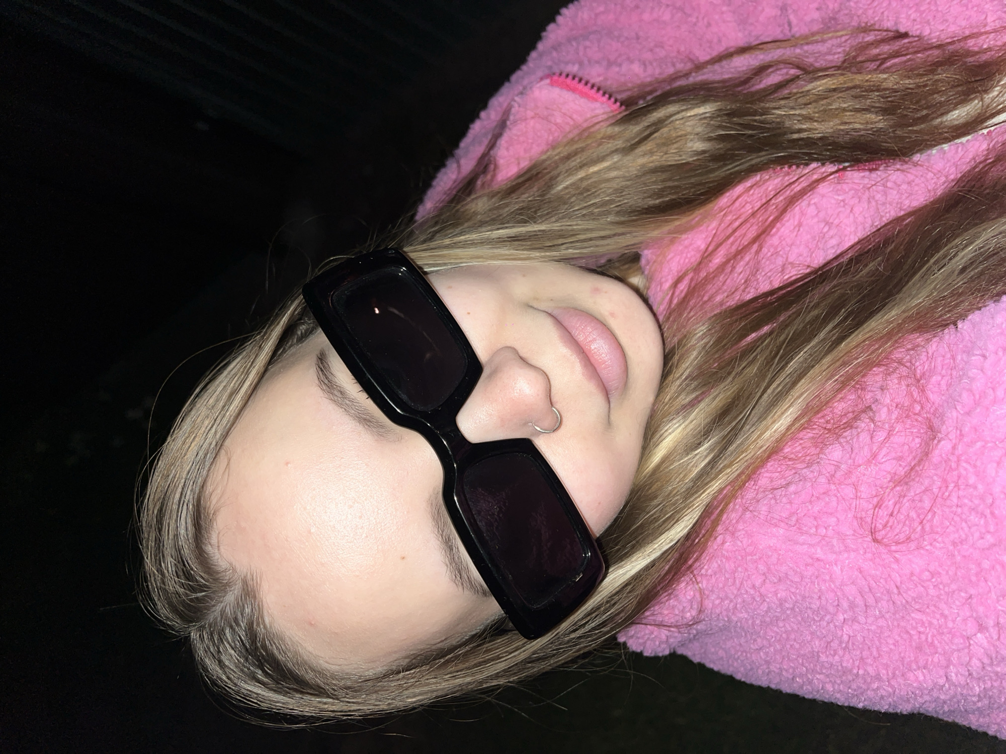 A young person wearing sunglasses and a pink jacket is lying down. The background is dark. The person has blonde hair and a nose ring. The photo may have been taken at night or in a dimly lit area. No elements point to Novopokrovskaya, Russia.