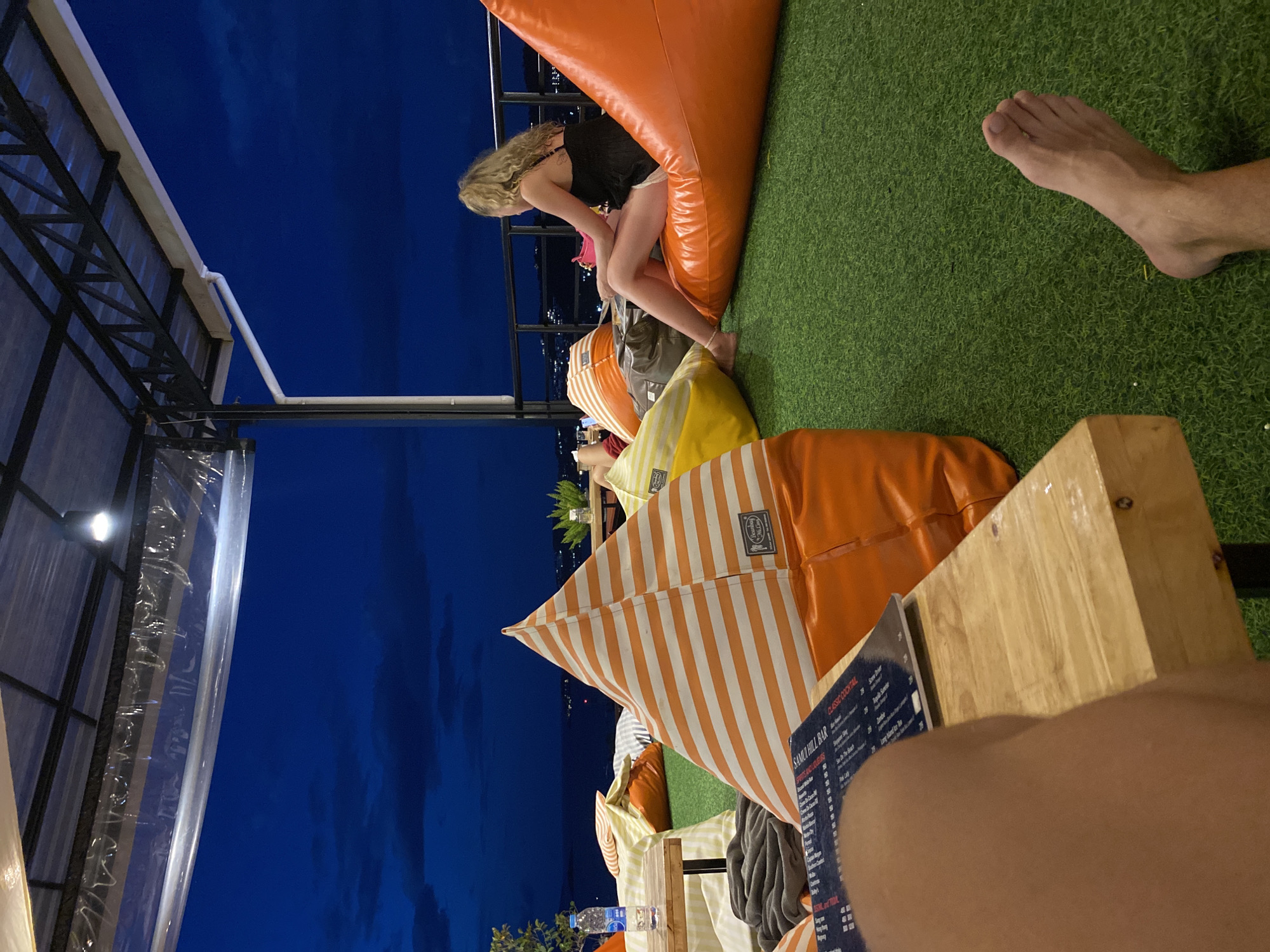 The image captures an outdoor setting at night, likely a rooftop bar in บ้านบางรักษ์, Thailand, given the menu visible reads "Samui Hill Bar." A woman with blonde curly hair is sitting on a large orange beanbag chair, leaning forward. She is wearing a dark top and light-colored bottoms, and has a pink object in her hands. Several other colorful beanbag chairs are scattered around, creating a casual seating area. A clear plastic canopy is visible on the left side of the frame, providing cover. The background shows a dark blue sky, suggesting the scene is taking place after sunset. In the foreground, the edge of a wooden table is visible, along with a person's leg and foot suggesting the image was taken from the perspective of someone sitting at the table. A menu from the bar is also on the table.