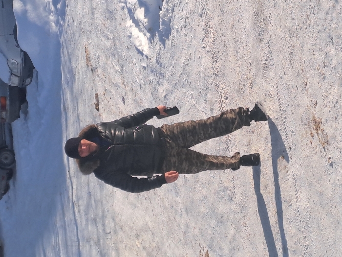 The scene, located in Bolshaya Elhovka, Russia, depicts an adult male walking on a snow-covered, cleared outdoor surface under bright sunlight. The man, positioned centrally, wears a black puffer jacket with a fur-lined hood, a black beanie, camouflage-patterned trousers, and dark boots. He holds a dark rectangular electronic device, consistent with a smartphone, in his right hand at waist level. Distinct tire tracks are visible on the icy and snow-covered ground. In the background, partially obscured by snow, are two parked vehicles: a light-colored sedan or hatchback and a darker vehicle. Long shadows are cast to the left of the subject, indicating a low sun angle. No other individuals or interactive subjects are present.
