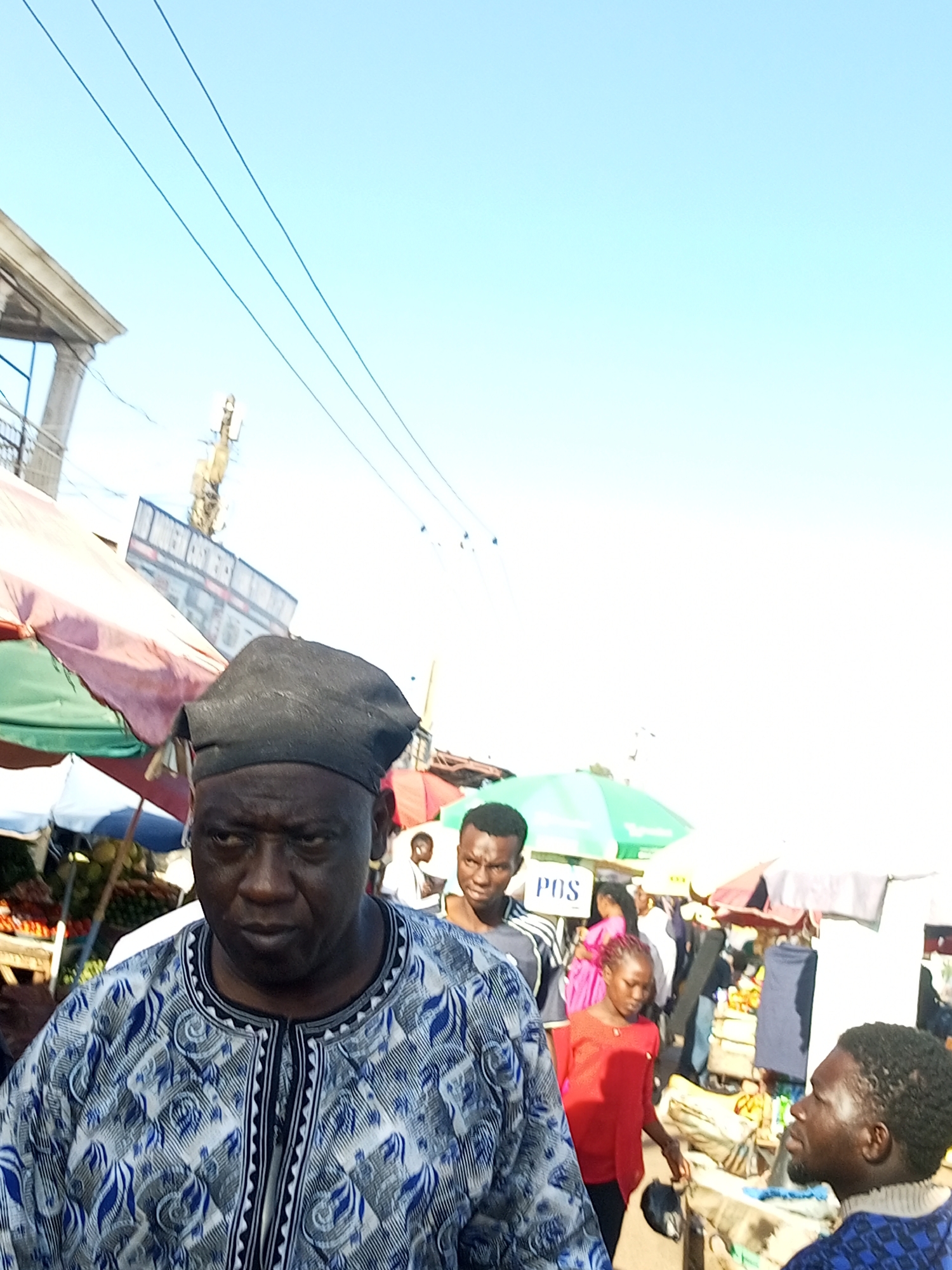 The image depicts a bustling outdoor market scene in Tempure, Nigeria. In the foreground, an older man wearing a black cap and a patterned blue and white robe is the central subject. He is looking to his left with a serious expression. Behind him, various market stalls with colorful umbrellas are visible. Other individuals are scattered throughout the market, including a younger man in a black and white striped shirt looking to the left, and a young woman in a red shirt also looking down. The background shows more market activity with stalls displaying goods and people interacting. The sky above is clear and blue. The overall impression is of a vibrant, everyday market day in a Nigerian city.