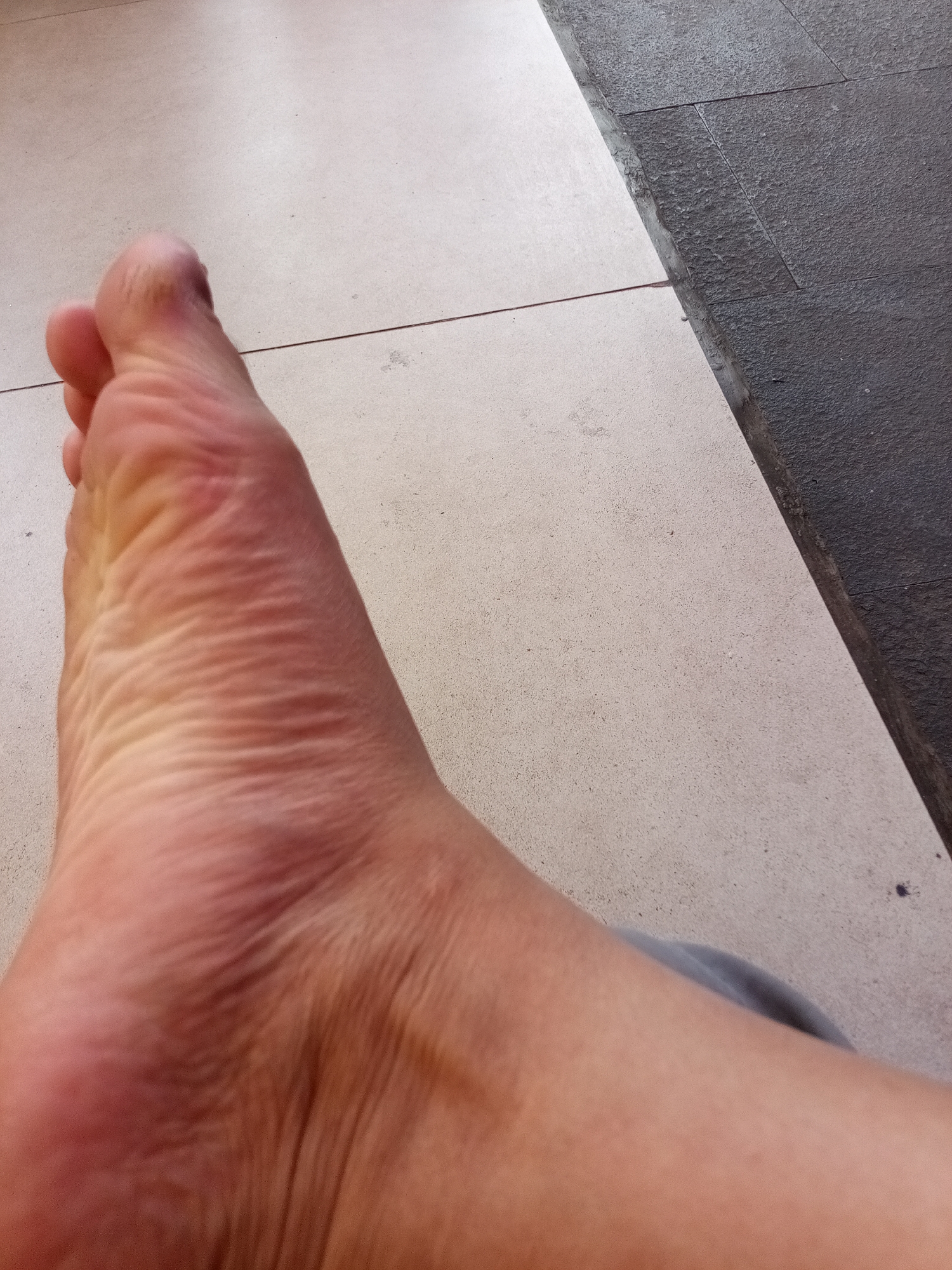 The image shows a close-up view of a person's left foot resting on light-colored tiled flooring. The foot is positioned diagonally, with the toes pointing towards the upper left corner. The skin of the foot appears to have some redness and wrinkles, particularly on the sole. To the right of the foot, the flooring transitions to darker, textured tiles, indicating a change in surface or an outdoor area. The lighting suggests it is daytime. The scene is devoid of any specific urban landmarks or identifiable features that would place it within Sariwangi, Indonesia.