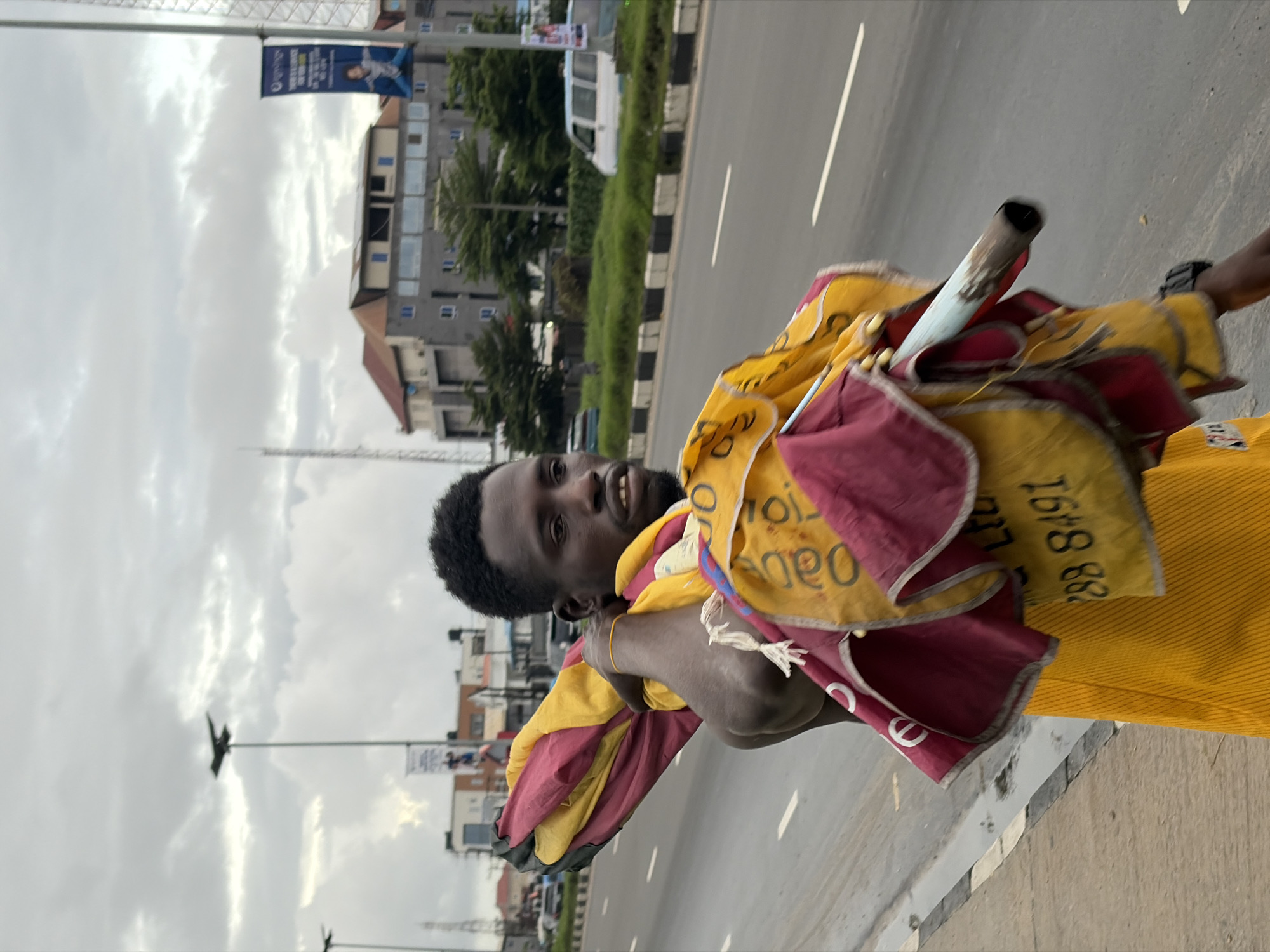 A man is walking on the side of a multi-lane road in Igboefon, Nigeria, with a folded umbrella slung over his shoulder. He is wearing a yellow sleeveless shirt. The umbrella is a vibrant yellow and maroon. In the background, there are buildings, streetlights, and vehicles on the road. The sky is overcast.