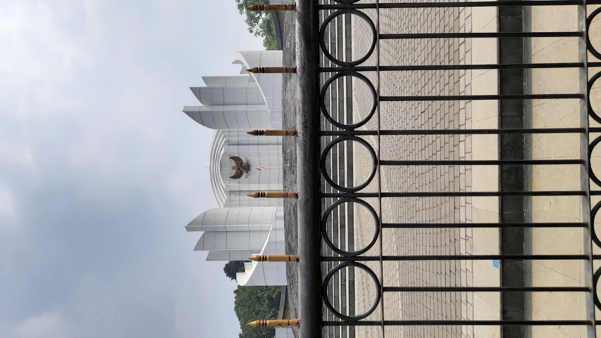 The image shows a modern white building with a curved facade and pointed architectural elements, resembling a stage or amphitheater. In the center of the building, a golden eagle emblem is displayed, and below it, a flag is visible. A set of stairs leads up to the building, and the foreground is occupied by a dark metal fence with circular patterns and decorative pointed finials. The ground in front of the building is paved with interlocking stones. Green trees are visible on the left side of the building. The sky is overcast with a few clouds. This building is likely the Monumen Perjuangan Rakyat Jawa Barat (West Java People's Struggle Monument) in Bandung, Indonesia.