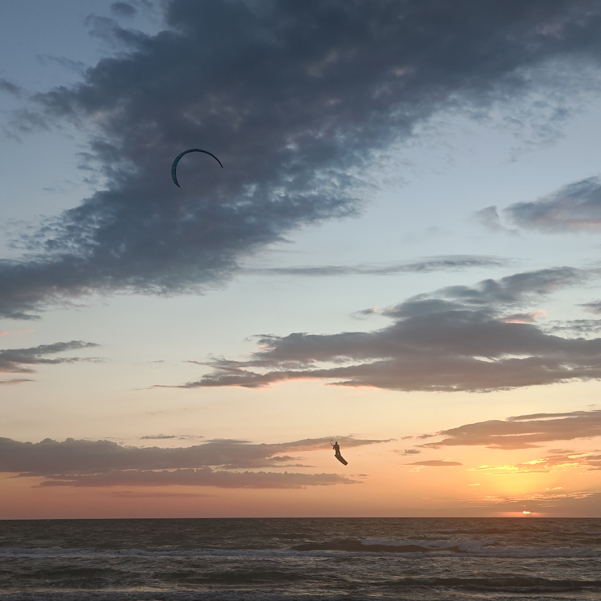 The image depicts a sunset over the ocean, with two kite surfers visible. One kite is in the upper left quadrant of the image, and the kite surfer is in the center-right quadrant. The sun is setting on the horizon, and there are waves breaking on the shore. The sky is cloudy, with a mix of dark and light clouds. The location is Unknown, Unknown.