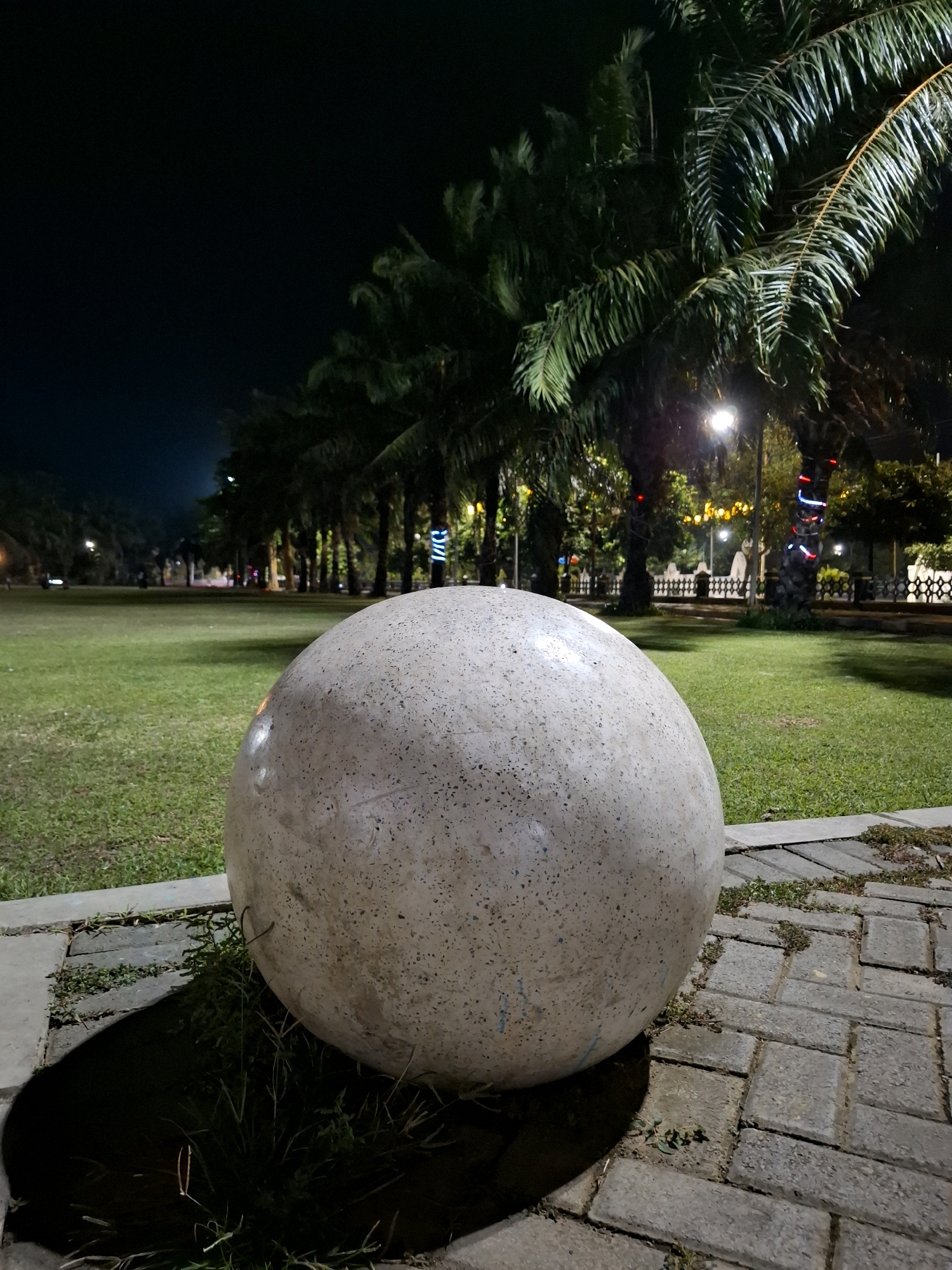 The image shows a large, speckled, spherical concrete sculpture in the foreground, positioned on a paved walkway. The walkway is made of rectangular pavers with grass growing in the cracks. Behind the sculpture is a well-maintained green lawn. A line of palm trees with dense foliage extends into the background, with some trees decorated with colorful lights. Streetlights are visible, illuminating the scene, and the sky is dark, indicating nighttime. In the distance, there appears to be a fence and some buildings. The location is identified as Pandean, Indonesia. There are no people or animals visible in the frame.