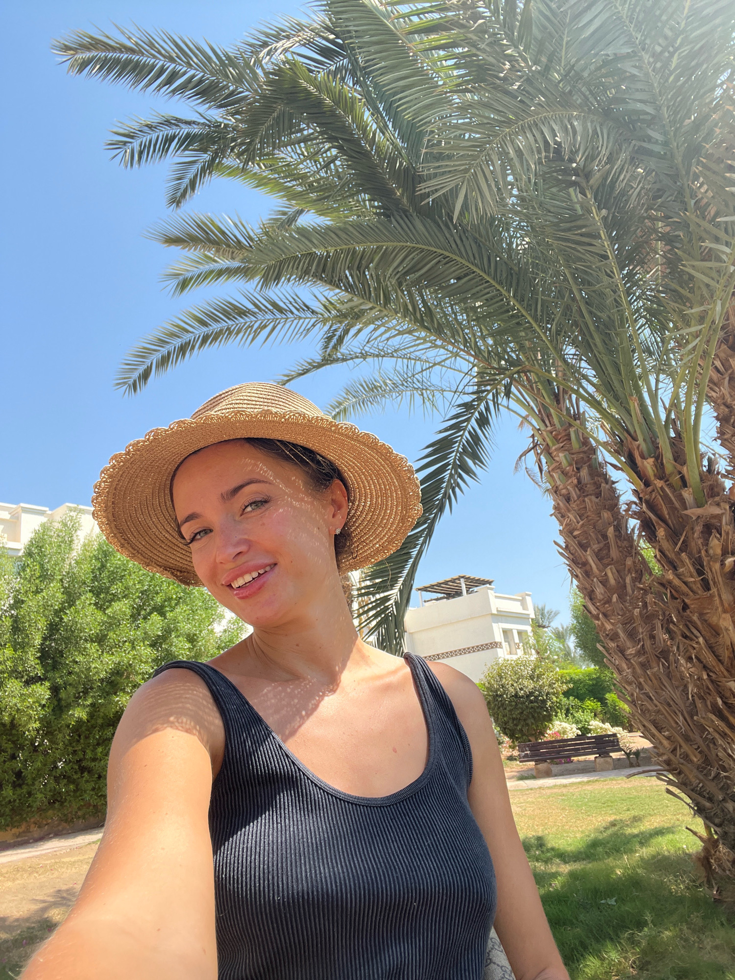 A woman wearing a black tank top and a straw hat takes a selfie outdoors. She is smiling and looking at the camera. She has green eyes and fair skin, with some freckles. Behind her, there is a palm tree and some buildings in the background. The sky is clear and blue. The scene appears to be a sunny day.