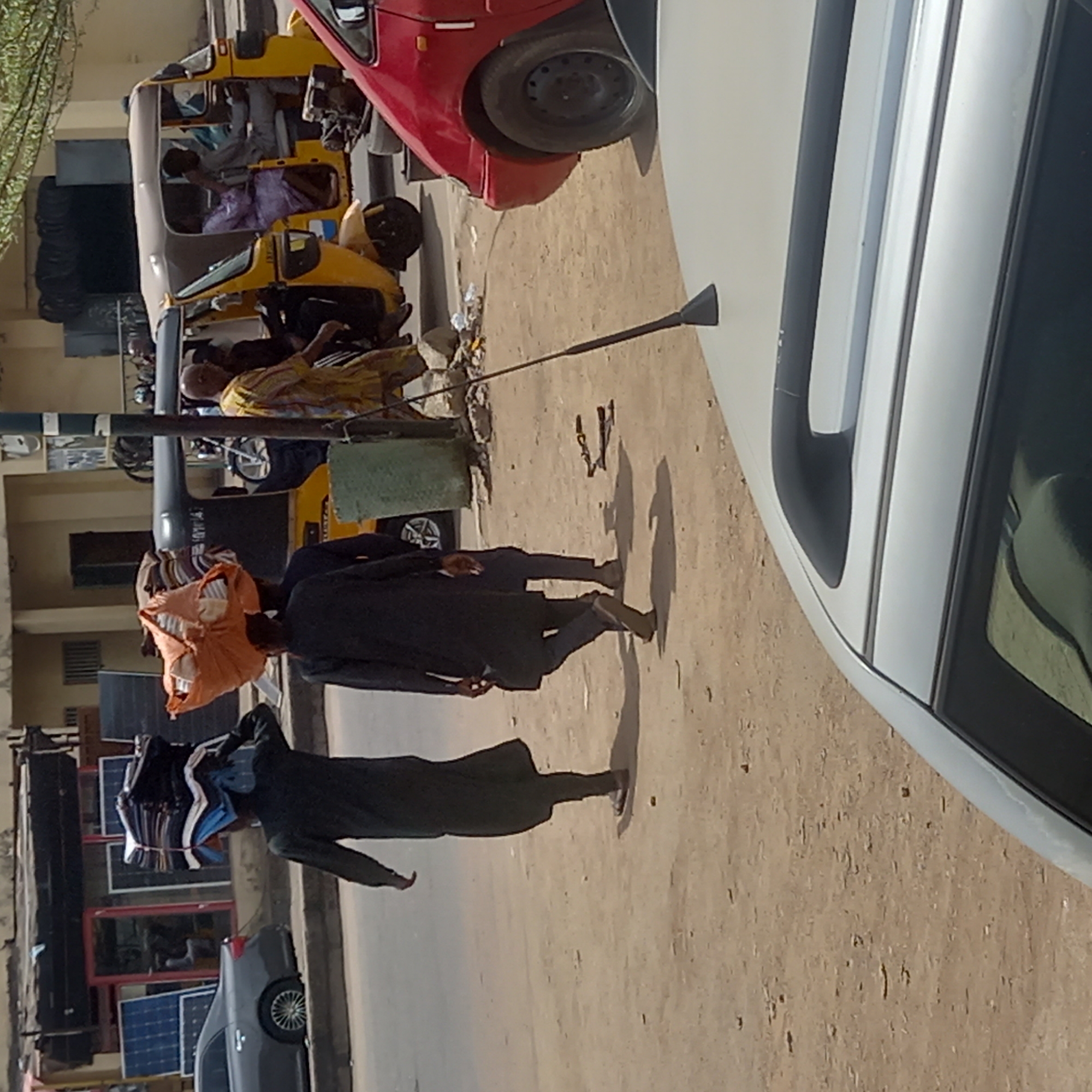 On a sunny day in Dadi Riba, Nigeria, a street scene unfolds. Two individuals are walking on a dirt road, each carrying items on their head. The first person has an orange cloth wrapped around their head, and the second is carrying a bundle of clothes. Various vehicles, including a red car, a yellow auto rickshaw, and a gray car, are parked along the road. Solar panels are visible near a building in the background. Other individuals are present, some inside the auto rickshaw. Trash is scattered on the ground. A portion of a white vehicle is visible on the right side of the image.