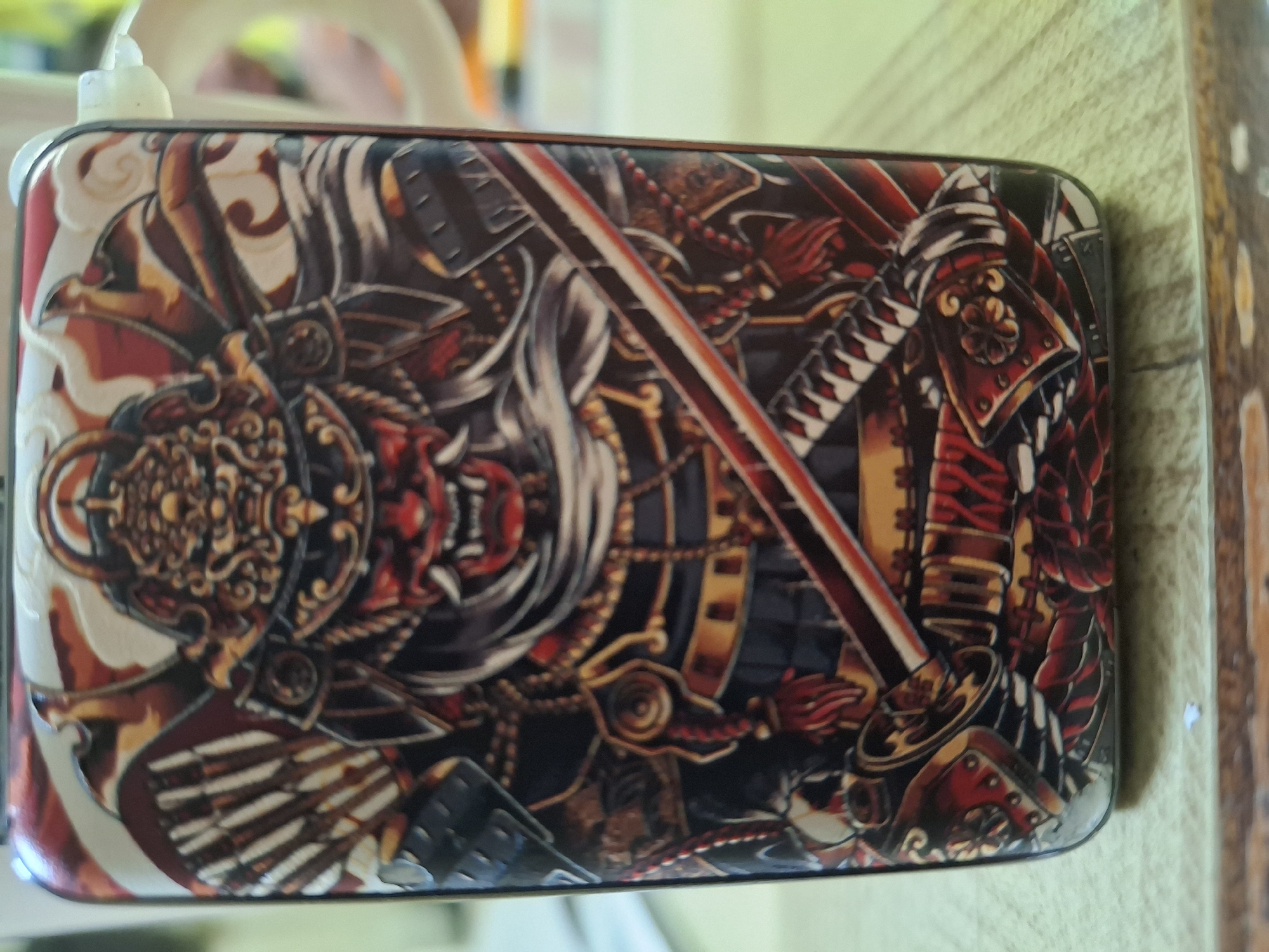 A close-up shot of a rectangular object, possibly a phone case or power bank, with an intricate graphic depicting a samurai warrior, complete with armor, a menacing mask, and a katana sword. The color palette predominantly features reds, blacks, golds, and whites, creating a visually rich and detailed illustration. The object rests on a surface, possibly a table, with a portion of what appears to be a wooden panel visible in the background. No details suggest a location in Eemshaven, Netherlands.
