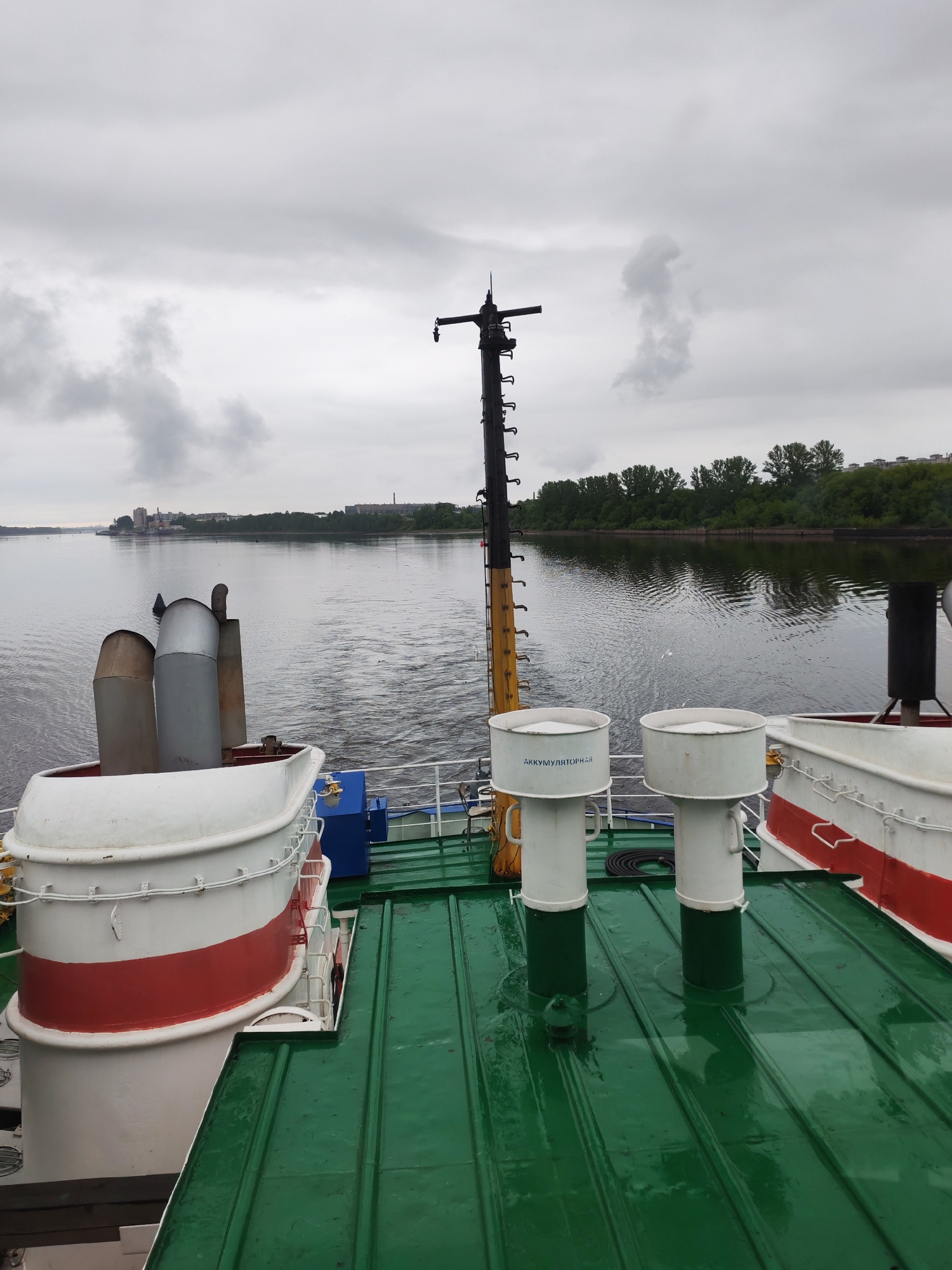 View from the deck of a ship sailing on a river near Bol'shie Mkhi, Russia. The scene is overcast. Visible on the deck are ventilation shafts, rescue boats, and a mast. In the background is a shoreline with buildings and trees.