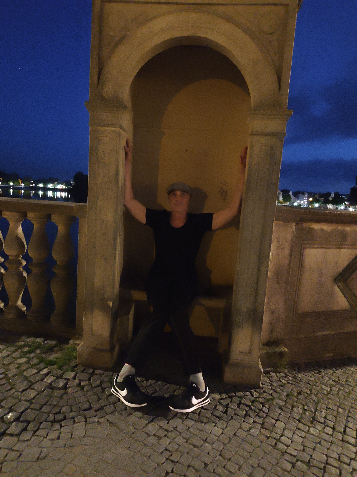 A man in a black t-shirt, black pants, a gray cap, and black Nike sneakers is posing with his arms spread and leaning on the stone pillars of an arched alcove. He is looking towards the camera. The background shows a body of water with lights reflecting on it, and buildings with lights in the distance. The ground is paved with cobblestones, and grass is growing in between some of the stones. The sky is a deep blue, indicating it is dusk or nighttime. The scene appears to be along a waterfront in Schwerin, Germany, possibly near the Schwerin Castle or along the Ziegelsee.