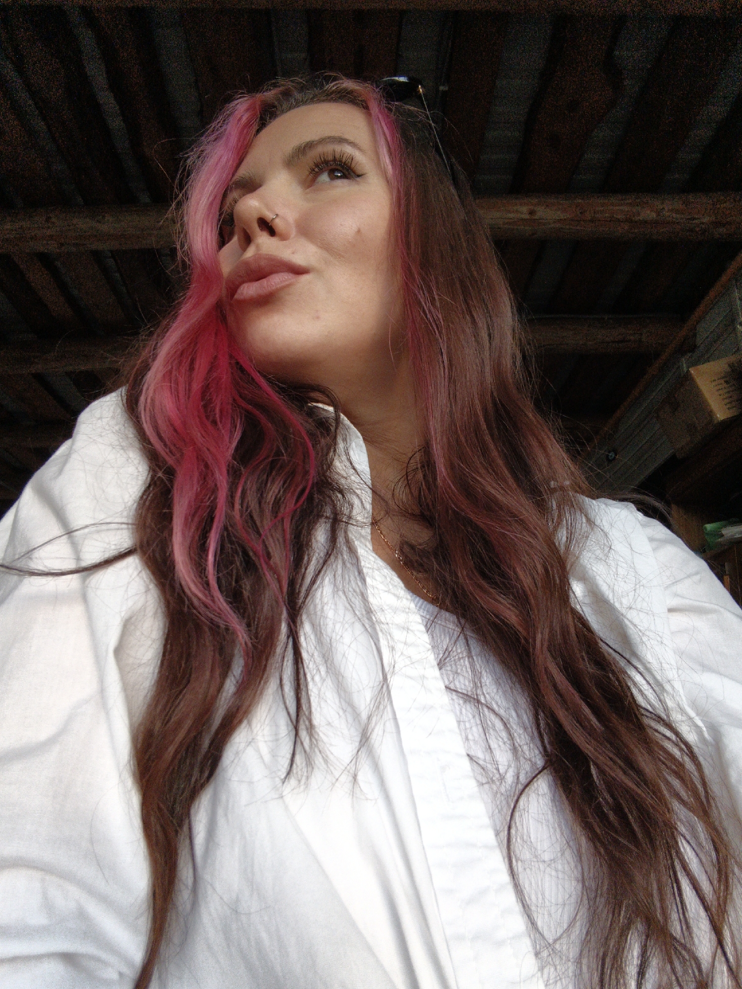 A close-up, low-angle shot shows a woman with long, dark brown hair streaked with bright pink, looking upwards and to her left. She is wearing a white collared shirt, and a delicate gold chain is visible around her neck. Her facial features are softly lit, and she has a nose ring. The background consists of dark, wooden beams, suggesting an indoor or covered outdoor setting, possibly a rustic structure. The location in the city of Ольховка, Russia, is not discernible from the visual information provided in the image itself.