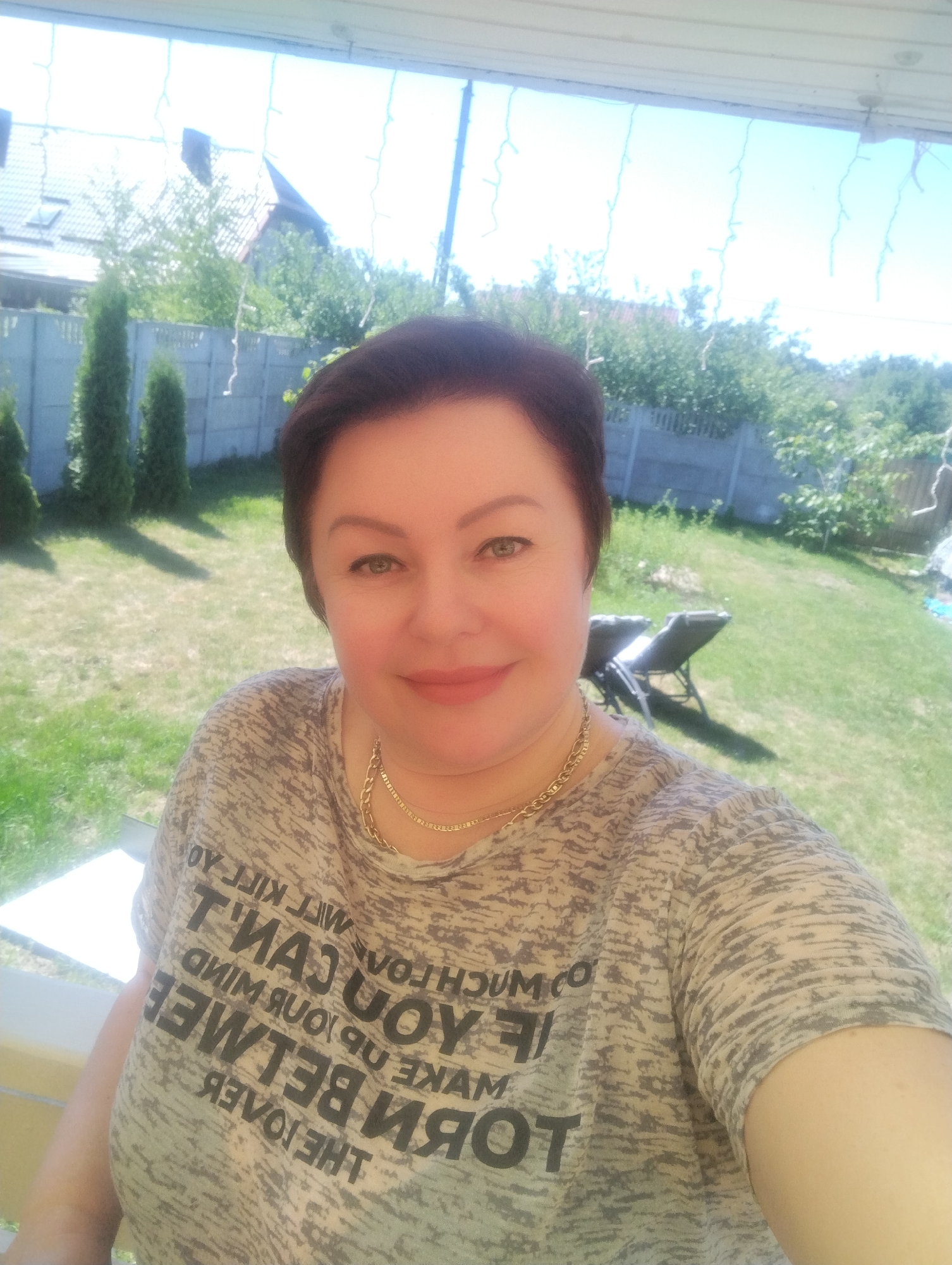 The image shows a woman with short, dark hair and light skin, wearing a grey, patterned t-shirt with a gold chain necklace. She is in a backyard setting, with a green lawn and trees visible in the background. There is a light-colored fence and some buildings in the distance. The sky is blue and clear. The woman is smiling and looking at the camera, suggesting a casual, outdoor moment. The location is identified as Svetly, Russia.