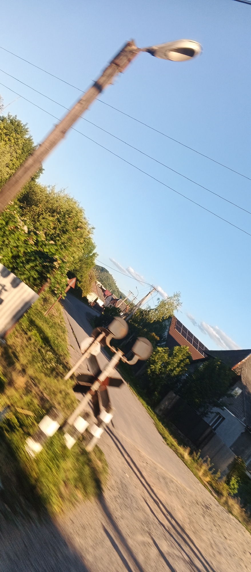The image, taken in Svaliava, Ukraine, captures a slightly tilted view of a rural road. The scene is dominated by a clear blue sky with a few wispy clouds. In the foreground, a weathered wooden pole with a streetlamp extends upwards and to the right, its details blurred due to motion. Several power lines are visible against the sky.
To the left, lush green trees and foliage line the roadside, partially obscuring a corrugated metal structure. A road crosses the frame, with a railway crossing signal featuring two circular lights and a red and white striped pole in the middle distance.
Further down the road, several houses are visible, some with dark tiled roofs. One house in the middle distance has solar panels installed on its roof. The road is paved and appears to be a residential street. The sunlight casts long shadows across the road, indicating it is likely late afternoon or early evening. The overall impression is a quiet, possibly rural, setting in Svaliava.
