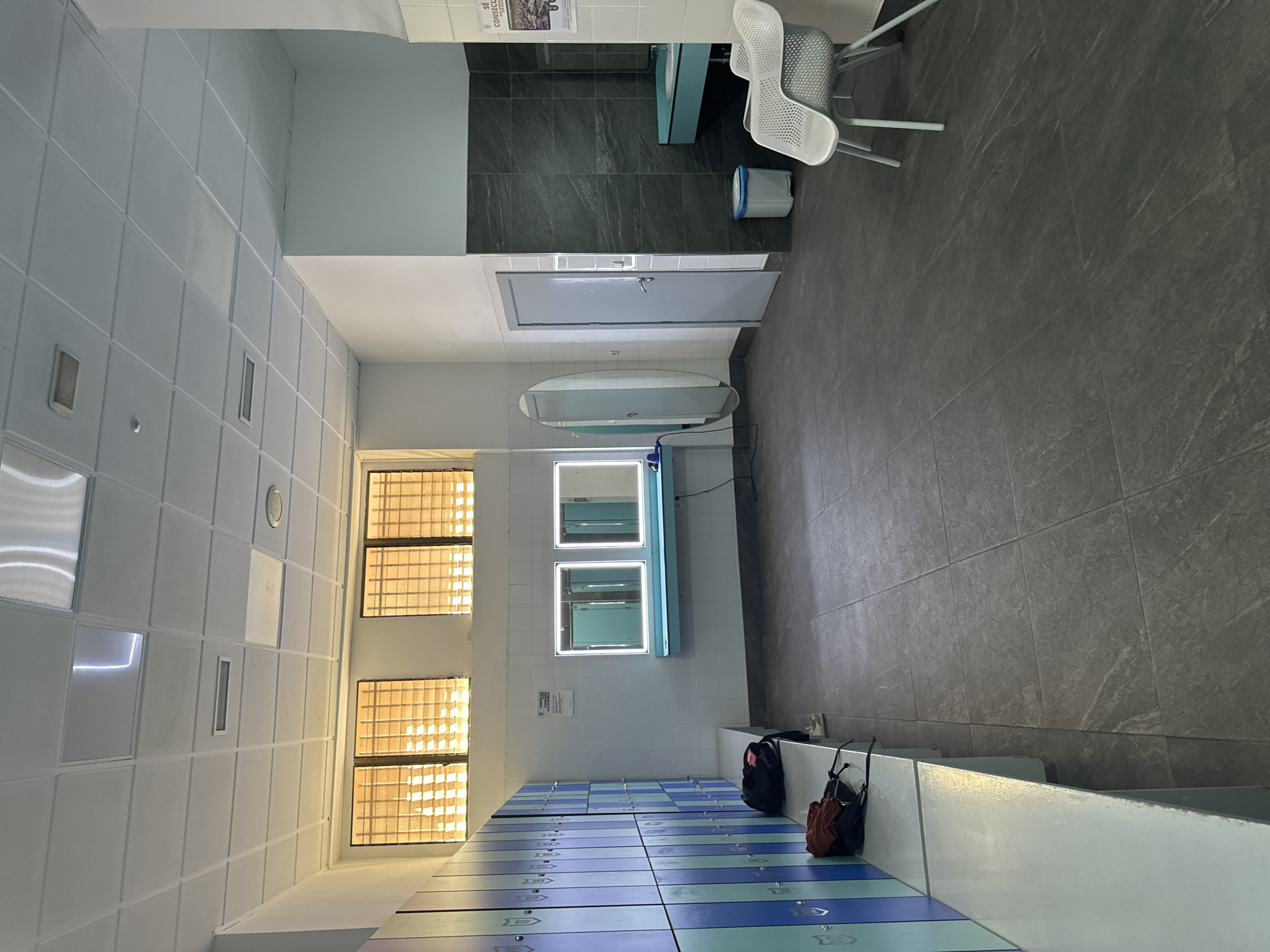 This media file shows a locker room in Murcia, Spain. The room is brightly lit, with fluorescent lights on the ceiling and illuminated mirrors above the lockers. The lockers are blue and white, with a row of them visible in the foreground. Two bags are placed on top of a bench near the lockers.
In the background, there are two white chairs and a trash can. A gray door is visible on the right side of the image. The floor is tiled in a dark gray color, and the walls are white with some gray tiles in the back. There are windows on the left side of the image that let in natural light.
The scene appears to be empty, with no people visible. The overall atmosphere is clean and functional, suggesting a public space like a gym or a public restroom. There is a sign on the wall near the mirrors, but the text is too small to read.