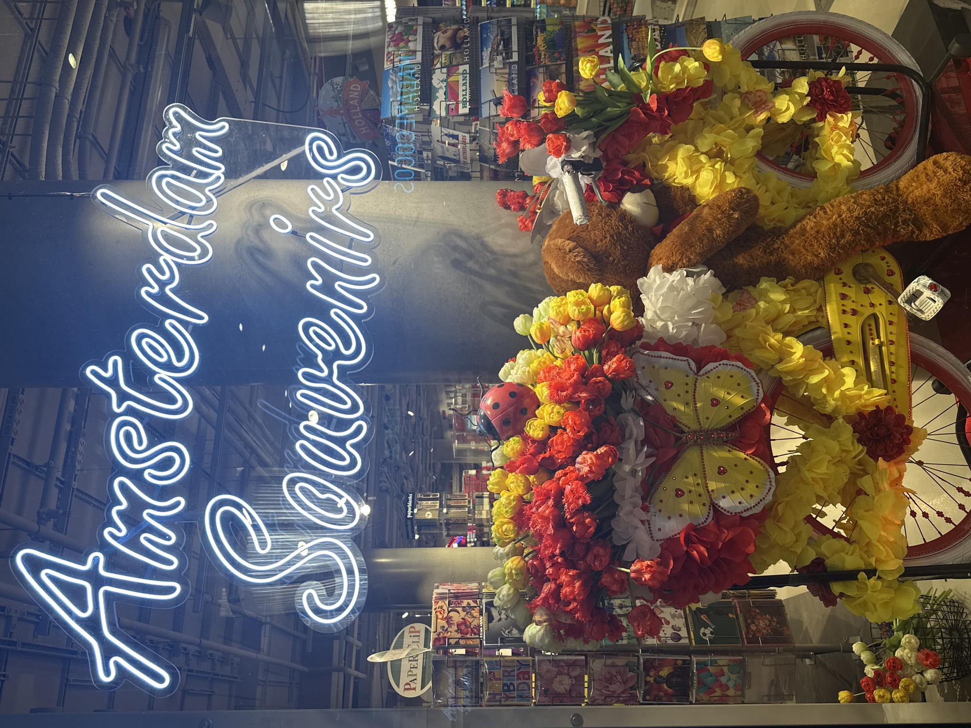 The image depicts a storefront window in Amsterdam, Netherlands, featuring a vibrant display for "Amsterdam Souvenirs." A neon blue sign, prominently placed at the top of the frame, spells out "Amsterdam Souvenirs" in a cursive font. Below the sign, a miniature yellow bicycle serves as the central element of the display. The bicycle is adorned with an abundance of colorful artificial flowers in shades of red, yellow, and white, creating a festive and eye-catching arrangement. A large, plush brown teddy bear is seated on the bicycle, leaning to its right. A large yellow butterfly with red accents is attached to the front of the bicycle. A ladybug decoration is placed among the flowers. Behind the bicycle display, shelves stocked with various souvenirs, including postcards and other Dutch-themed items, are visible through the glass. The background shows the interior of a shop with lighting and additional merchandise. The overall scene is designed to attract tourists and promote local products.