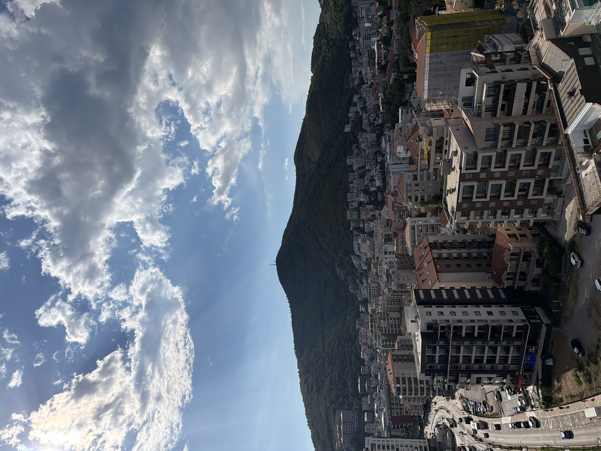An aerial view captures a dense urban landscape in Budva, Montenegro, with a prominent, tree-covered mountain dominating the background under a partly cloudy sky. The foreground is filled with a variety of multi-story buildings, ranging in architectural style and color. Some buildings feature modern designs with dark facades and balconies, while others display traditional red-tiled roofs. A busy street runs through the lower left of the frame, lined with parked cars and a few pedestrians. The mountain in the distance is covered in lush green vegetation and has a small structure, likely a transmission tower, near its summit. The sky above is a mix of blue patches and dramatic white and gray clouds, with sunlight breaking through in some areas, creating rays of light. The overall scene depicts a bustling coastal city nestled against a natural mountainous backdrop.