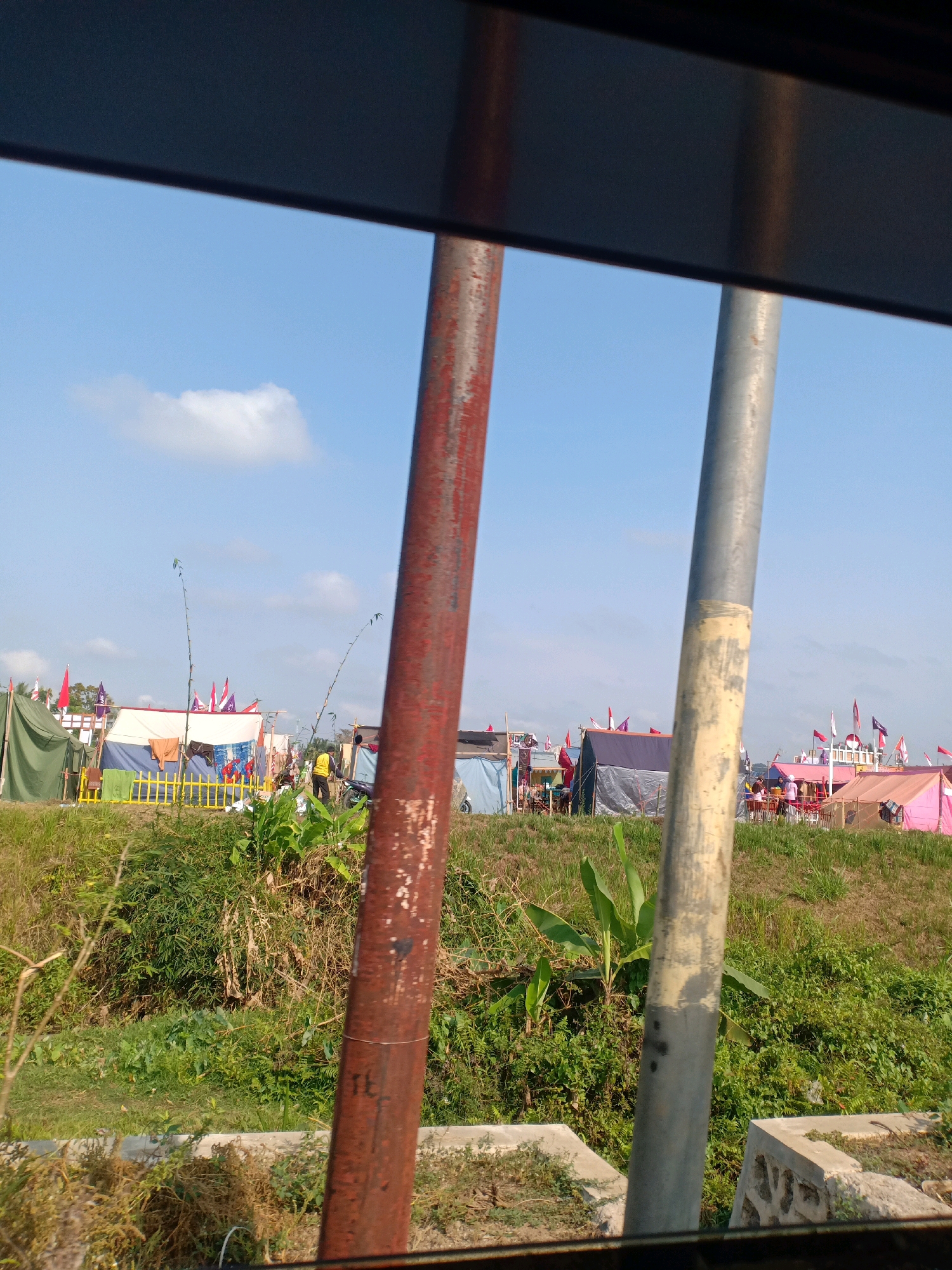The image depicts an outdoor scene with a clear blue sky and scattered clouds. In the foreground, two vertical poles, one rusted red and the other a lighter gray, are positioned prominently. Beyond the poles, a grassy embankment slopes upwards, leading to a collection of tents and temporary structures. These structures appear to be set up in a field, possibly for an event or festival. Several flags, predominantly red and white, are visible flying above the tents, suggesting a national or celebratory occasion.
There are a few individuals present, including one person wearing a yellow vest standing near a motorcycle. The overall impression is one of a gathering or campsite in a semi-rural or open area, likely in Tugusari, Indonesia, given the context. The environment is predominantly green with vegetation covering the ground and the embankment.
