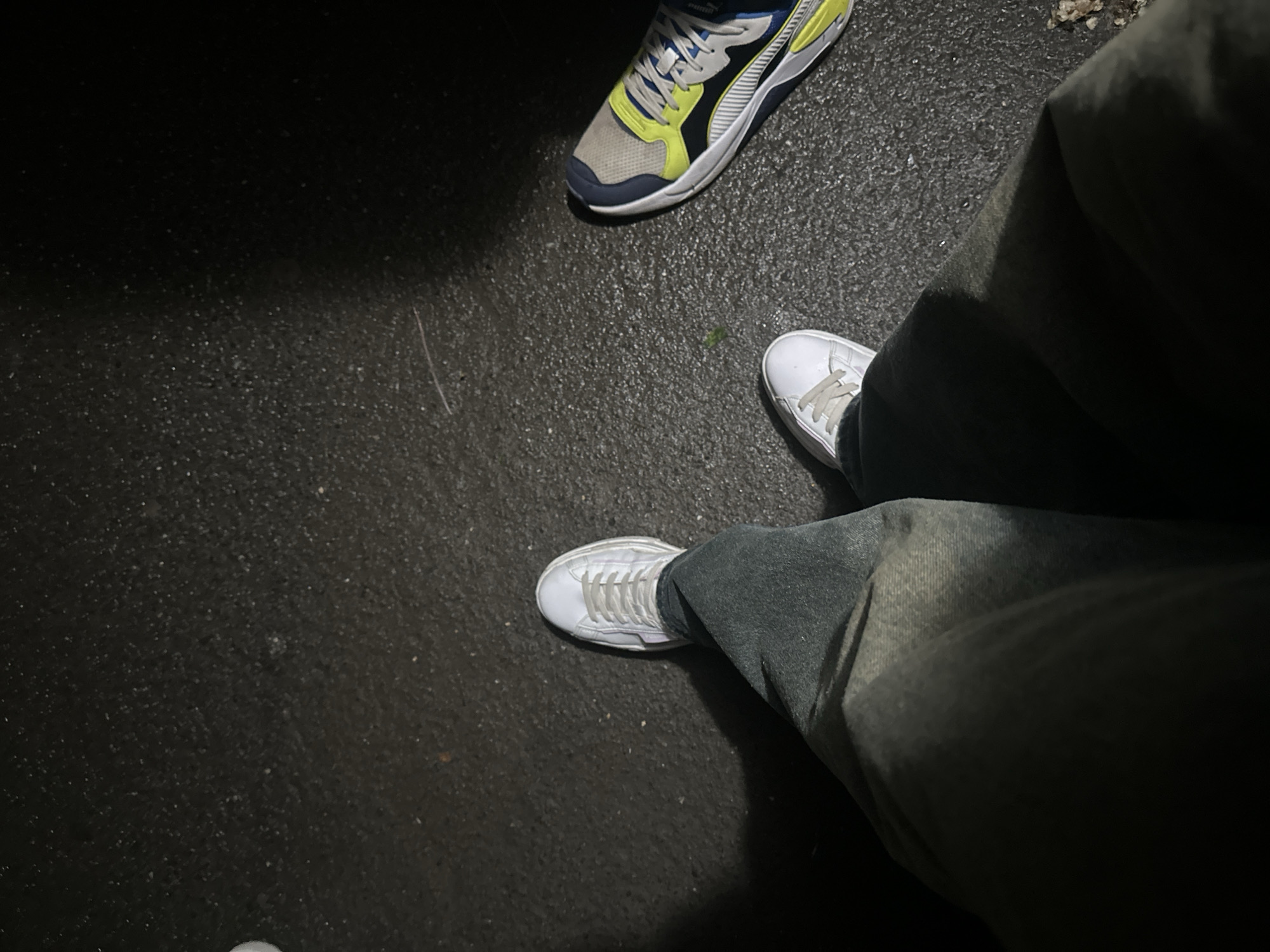 The image shows a low-angle view of a person's legs and feet standing on a dark, wet asphalt surface, likely a sidewalk or road. The person is wearing dark-colored pants and two different types of sneakers. On the left, a white sneaker is visible, and on the right, a multi-colored sneaker with yellow, blue, and grey accents is present. The lighting suggests it is nighttime, with a spotlight illuminating the area around the feet, casting shadows. The background is dark and out of focus. The scene is likely a public outdoor area in Togliatti, Russia, given the context provided.