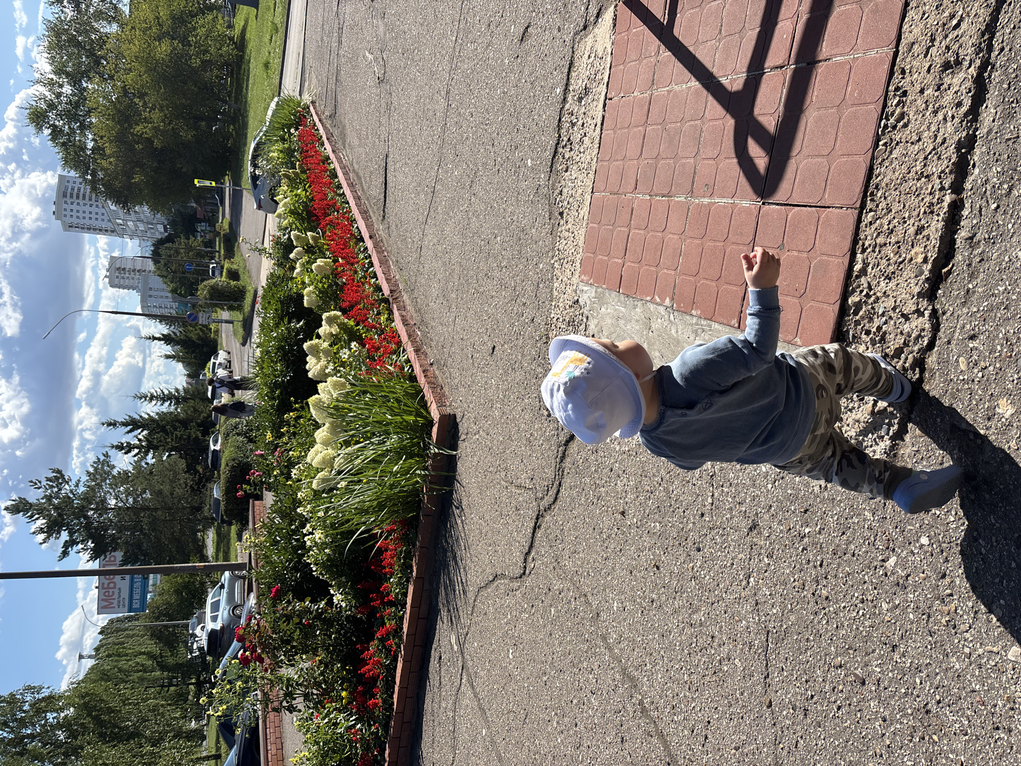 A young child wearing a blue shirt, camouflage pants, and a white sun hat walks on a cracked asphalt path in Naberezhnye Chelny, Russia. The child is facing away from the camera, towards a flower bed filled with red and white flowers. In the background, there are trees, apartment buildings, cars, and a billboard that reads "МЕБЕЛЬНЫЙ ЦЕНТР" (Furniture Center). The sky is blue with white clouds.