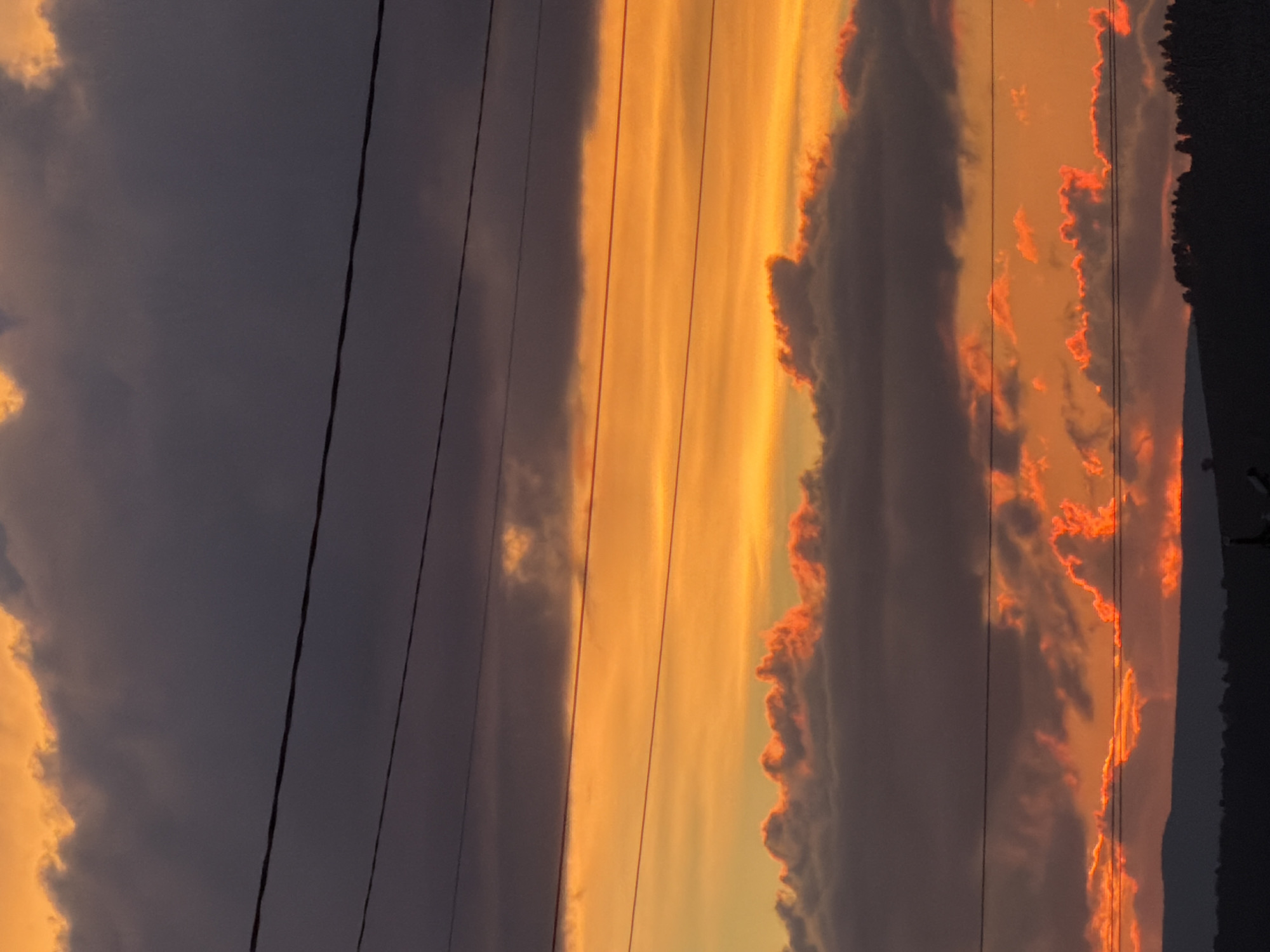 The image shows a dramatic sunset over a silhouetted landscape. The sky is filled with vibrant orange and yellow hues, with dark, moody clouds drifting across. Several power lines are visible, stretching horizontally across the frame, adding a man-made element to the natural scene. The lower portion of the image is dominated by the dark outline of hills and trees, creating a stark contrast with the fiery sky. The overall impression is one of atmospheric beauty and a sense of vastness.