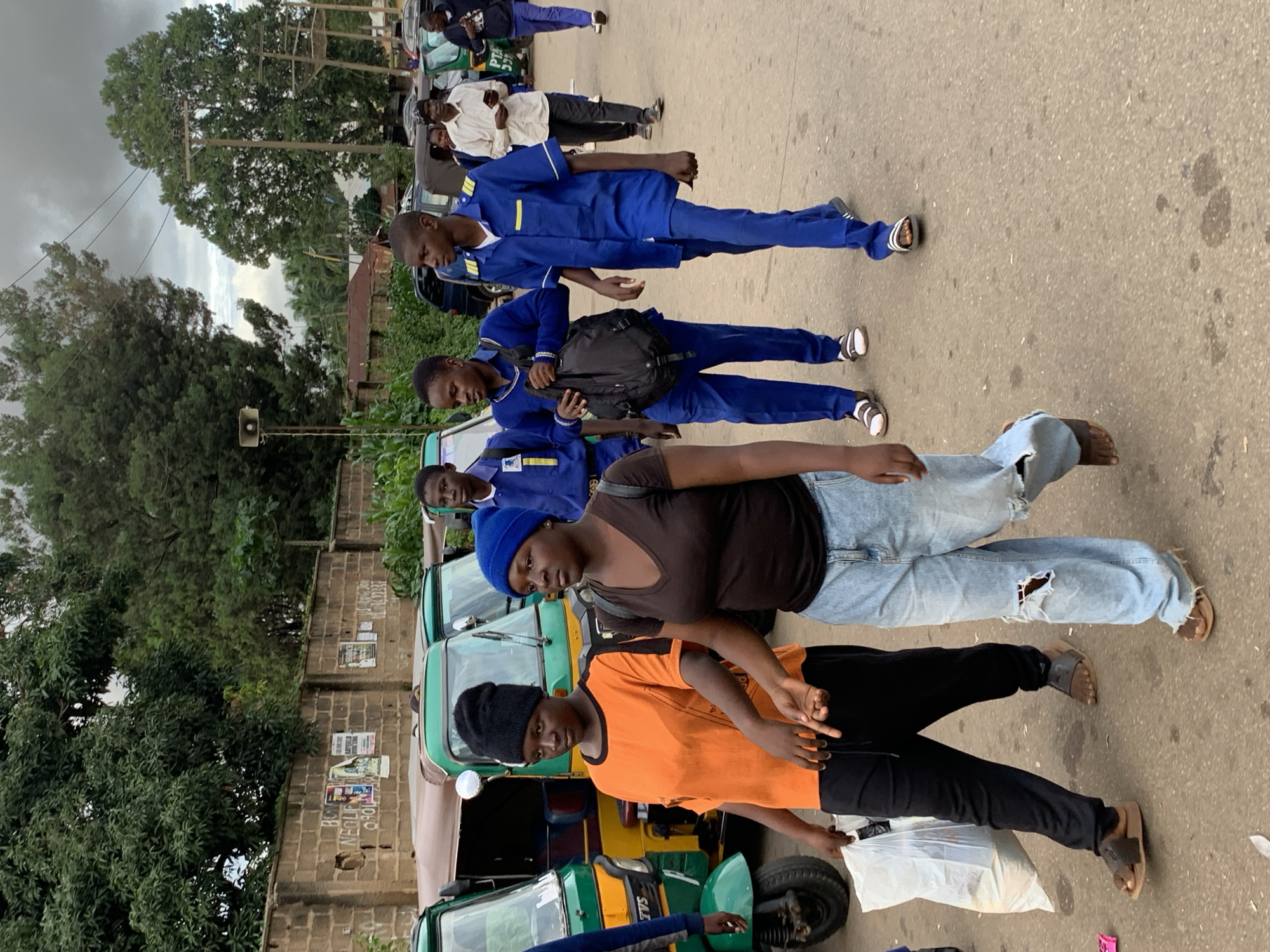 In Barakin Lerawi, Nigeria, a group of individuals walk along a paved road lined with trees and a brick wall. In the foreground, two young women walk together; one wears an orange t-shirt and a black headscarf, carrying a white plastic bag in her left hand. The other woman wears a dark brown v-neck shirt and ripped blue jeans, with a blue beanie on her head. Behind them, several young men in blue uniforms walk; some carry backpacks. In the background, several three-wheeled auto-rickshaws are parked. The sky is overcast, suggesting a cloudy day.