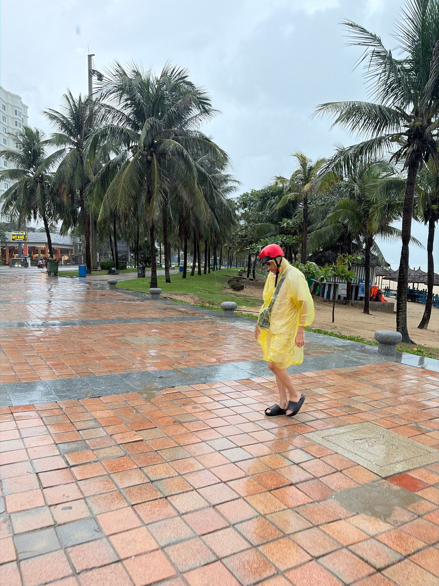 A person wearing a yellow raincoat and red helmet walks on a wet tiled pathway lined with palm trees. The scene appears to be a beach promenade in Đà Nẵng, Vietnam, as indicated by the presence of palm trees, beach huts with thatched roofs, and the cloudy sky suggesting potentially rainy weather. The pathway is made of reddish-brown square tiles with darker gray borders, and puddles of water reflect the overcast sky. In the background, a multi-story building and shops are visible, further suggesting an urban coastal location. The person is walking from left to right and is wearing dark sandals. The overall impression is a slightly gloomy but scenic day in a tropical city.