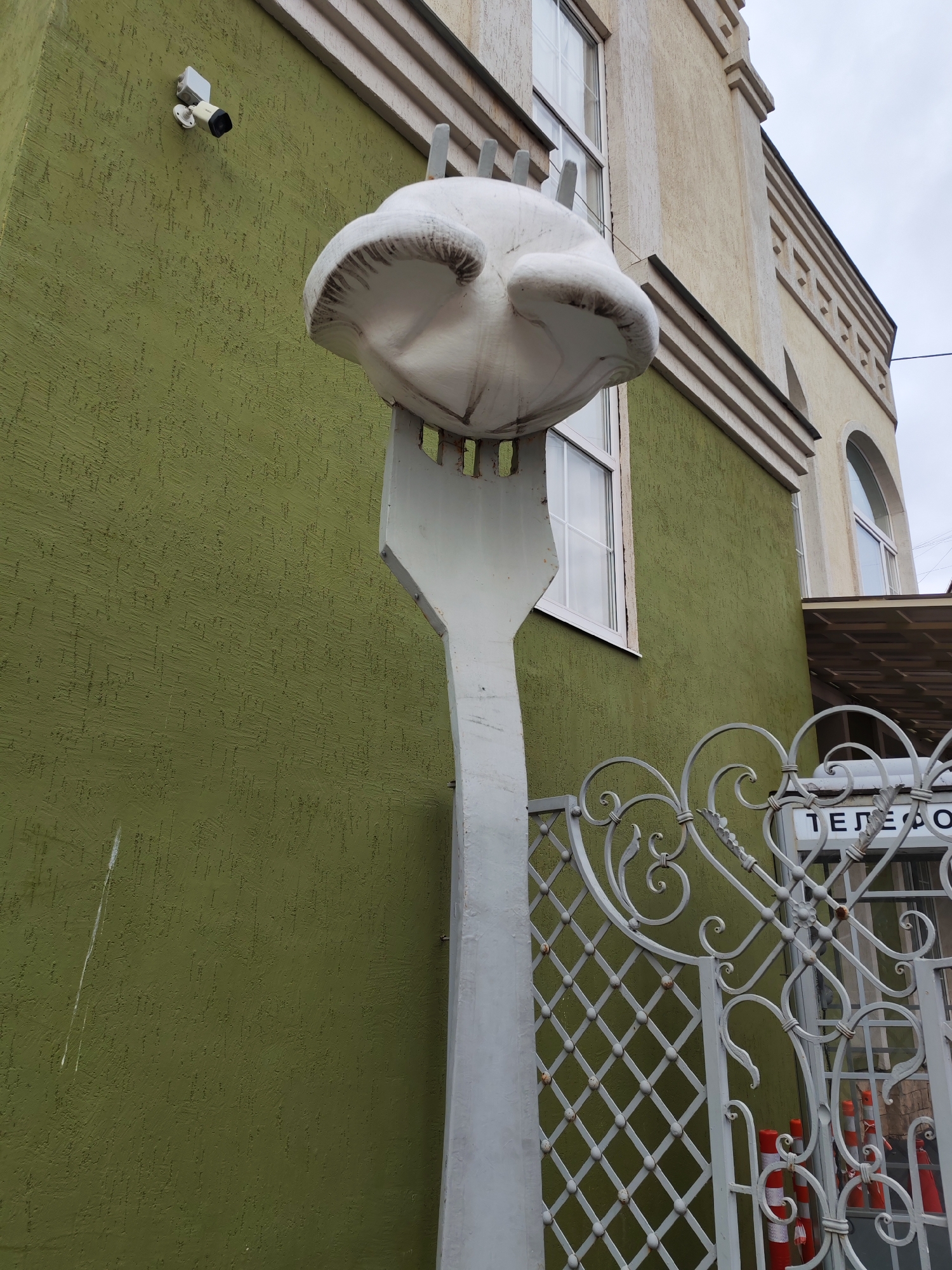A large, possibly sculptural, white garlic bulb impaled on a white metal fork stands adjacent to a building in Izhevsk, Russia. The building has both green and beige sections, with windows visible on both. A surveillance camera is mounted on the green section. In the foreground, a decorative metal fence obscures some of the scene. The word "Telephone" is partially visible in Cyrillic script on the fence.