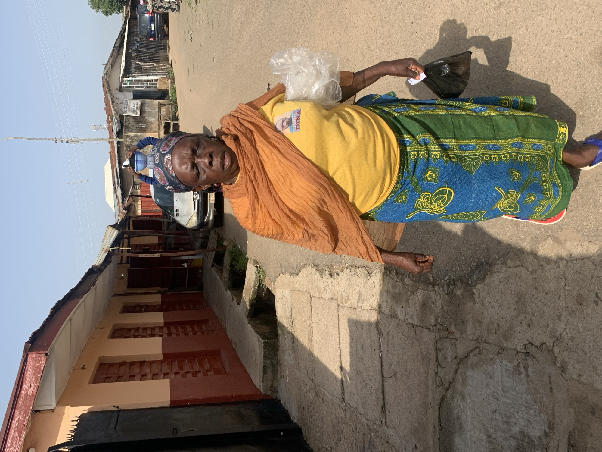 In Jos, Nigeria, on a sunlit day, an elderly Black woman walks down a paved street lined with buildings. She wears a yellow t-shirt with a picture of a man labeled "TANSIR DINA" on it, an orange headscarf, and a blue and green patterned wrap skirt. She carries a blue plastic basin on her head, a plastic bag filled with white material in her right hand, and a black plastic bag in her left. The street has a concrete sidewalk with a drainage ditch. In the background, several cars and buildings with corrugated metal roofs are visible.