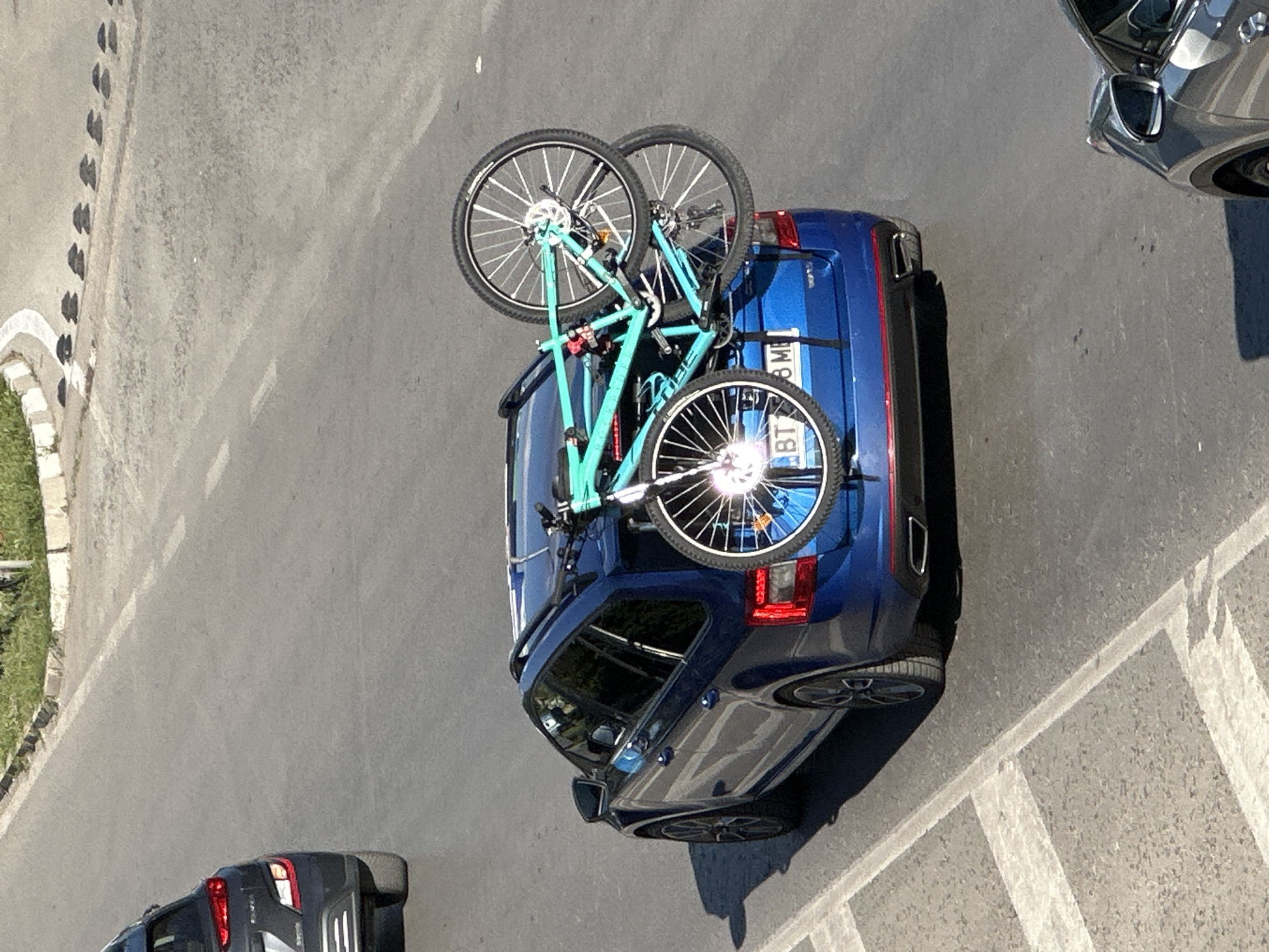 A blue station wagon is parked on a road in Veliko Tarnovo, Bulgaria, with two bicycles mounted on a rack on its rear. The car has a license plate that reads "BT 8 MB". The road is paved and has white lane markings. In the background, there is a sidewalk with a curb and some greenery. To the left of the blue car, a black SUV is partially visible. To the right, the corner of a silver car is visible. The scene appears to be captured on a sunny day.