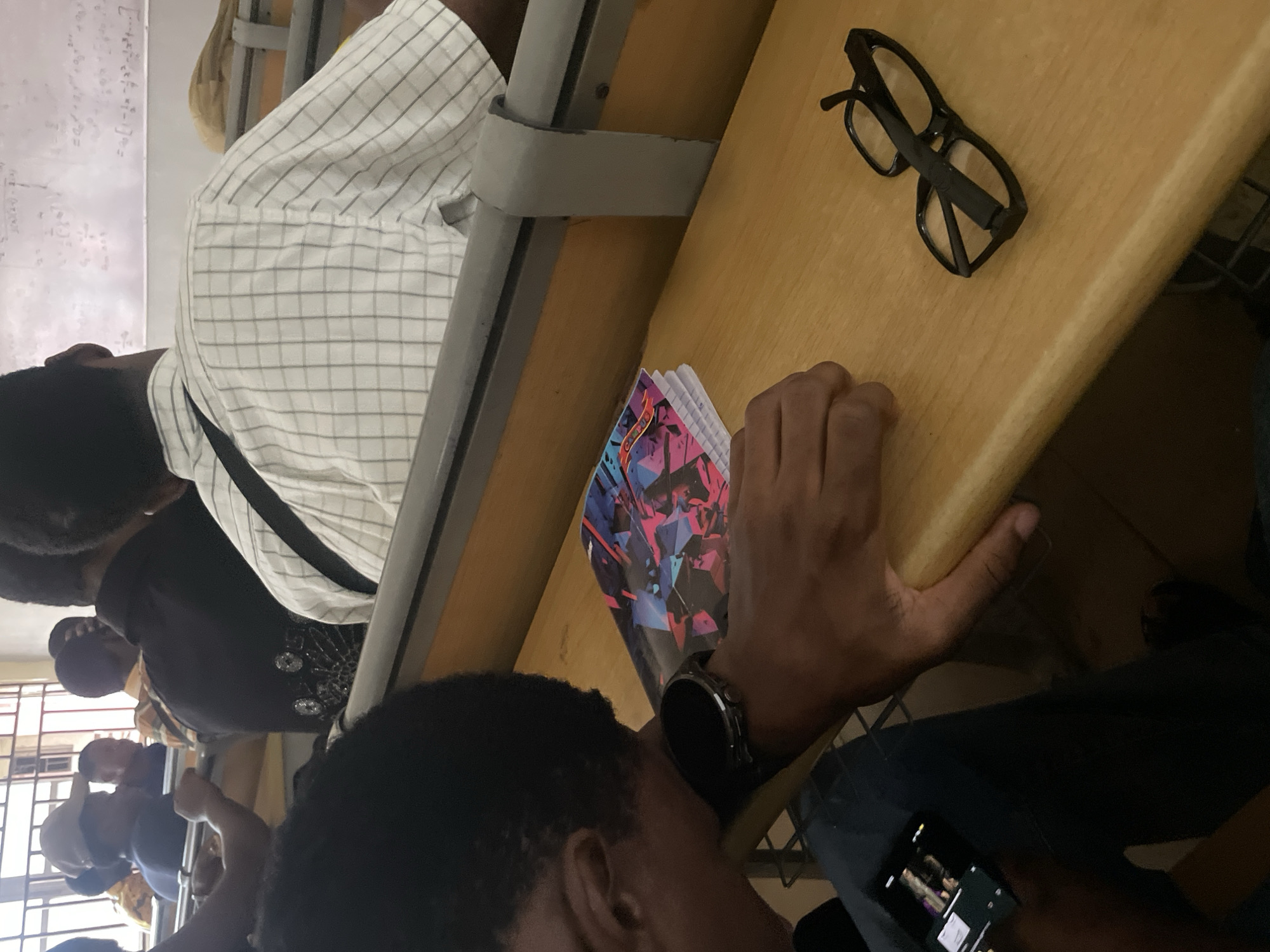 The image shows a classroom setting with several individuals seated at wooden desks. The focus is on a person in the foreground wearing a watch and holding a smartphone. In front of them are a notebook with a geometric design and a pair of black-rimmed glasses. The classroom appears to be in a state of partial activity, with students facing forward, suggesting a lecture or class is in progress. Mathematical equations are visible on a whiteboard in the background, indicating an academic environment. The location is specified as Ihiagwa, Nigeria.