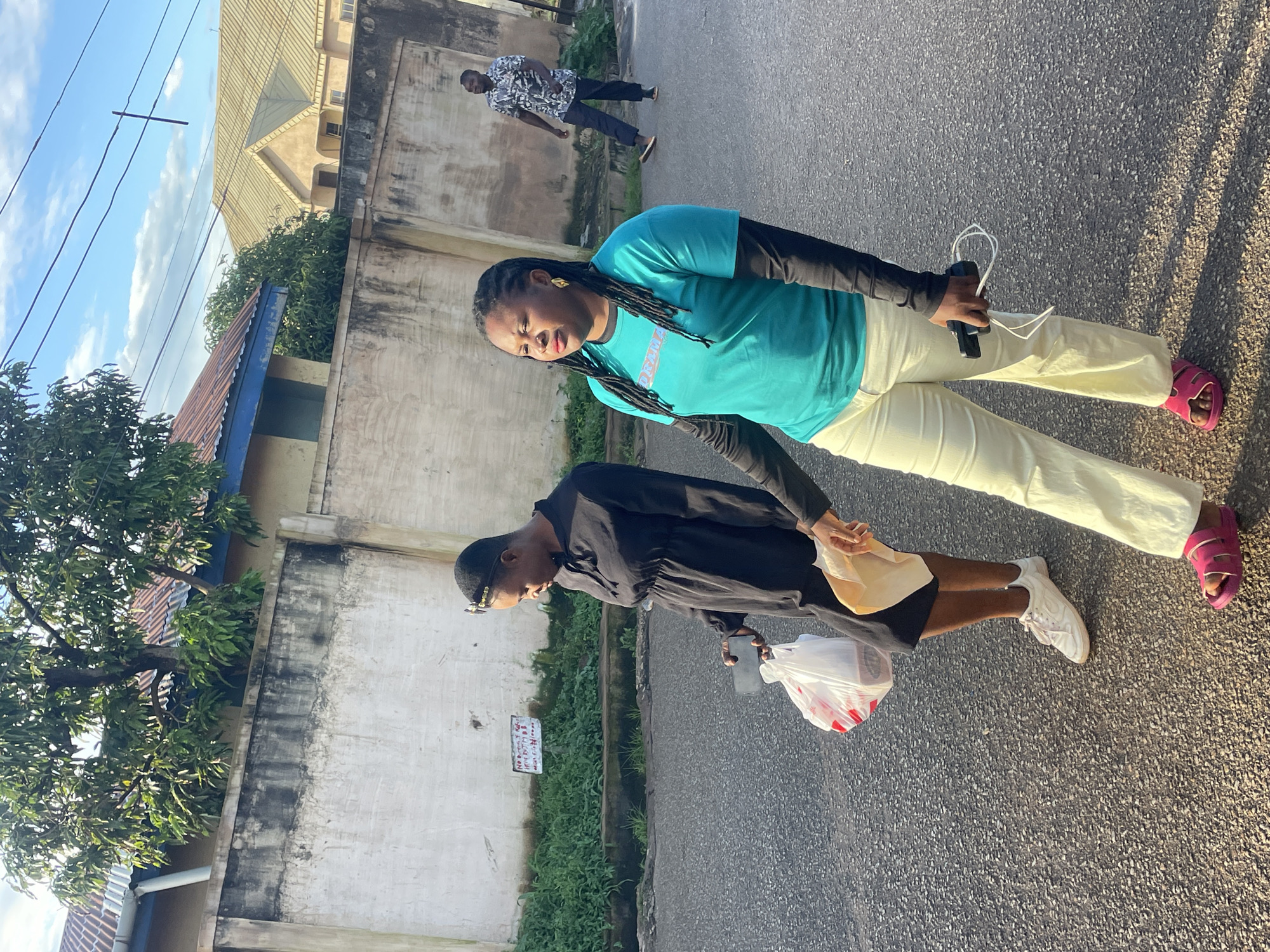 In Jos, Nigeria, two women walk on a paved road. The woman in the foreground is wearing a turquoise long-sleeved t-shirt, cream-colored pants, and pink sandals. She holds a phone in her right hand. To her left, another woman in a black dress and white sneakers walks, carrying a white plastic bag and a brown paper bag. In the background, a man in a patterned shirt and dark pants walks away from the camera. The scene is framed by a textured wall and lush green foliage, with buildings and power lines visible in the distance under a bright, partly cloudy sky. The lighting suggests it is late afternoon or early morning.