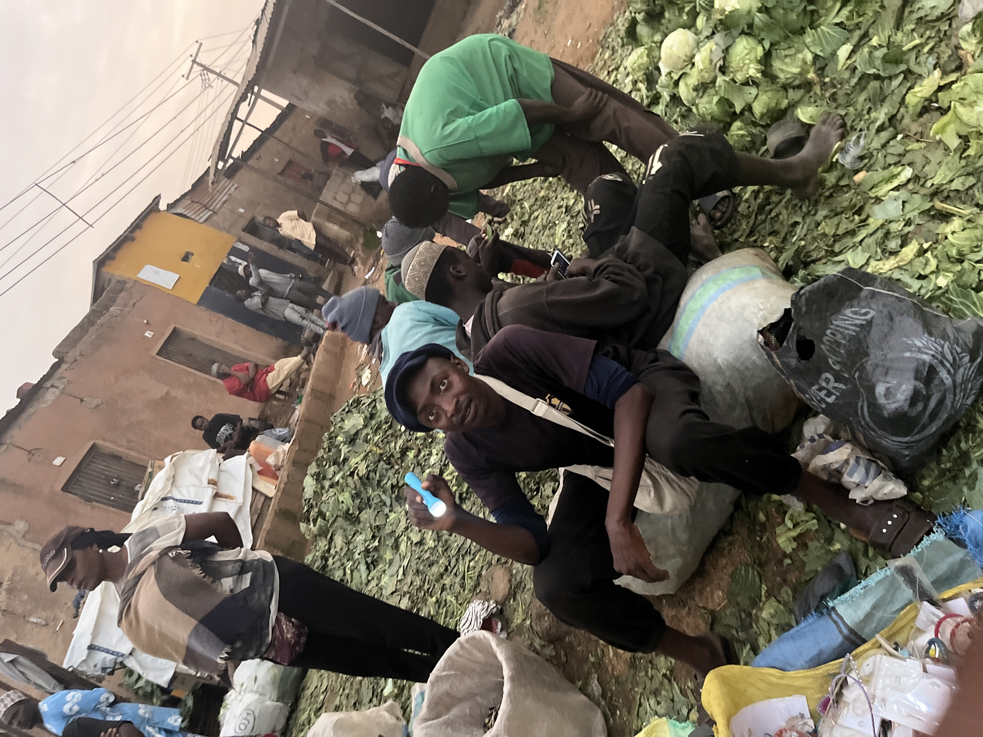 The image depicts a bustling outdoor market scene in Gura Top, Nigeria. In the foreground, a man in a dark t-shirt and cap sits on a large sack, holding a bright blue flashlight. He is surrounded by piles of leafy green vegetables, likely harvested produce. To his left, a large white bin contains what appears to be more produce or supplies. Behind him, other individuals are visible, some standing and others seated amidst the market stalls.
A woman wearing a baseball cap and a scarf draped over her shoulders stands to the left, looking away from the camera. She is dressed in dark pants and a white top. In the background, a row of buildings, some with corrugated metal roofs, lines the market area. Power lines crisscross the sky above the structures, indicating an urban or semi-urban setting. The ground is uneven and appears to be made of dirt or gravel, covered in scattered leaves and debris. Several large bags, some seemingly filled with produce and others with unknown contents, are scattered throughout the scene. The overall atmosphere suggests a busy commercial environment where goods are being sold and transactions are taking place.