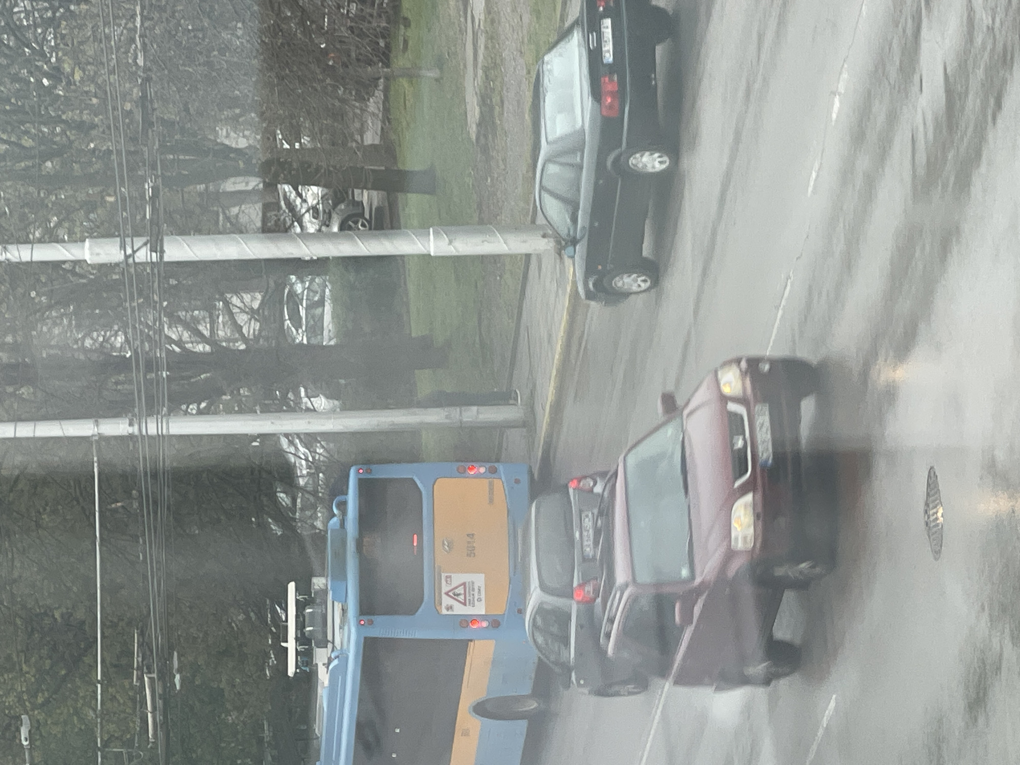 The image, taken from inside a vehicle looking out, depicts a street scene in Sofia, Bulgaria, on what appears to be a wet or recently wet day, indicated by streaks on the viewing surface and reflections on the road. The time of day is daytime, with overcast conditions.
In the foreground, a red delivery van, possibly a Mercedes-Benz Sprinter or similar model, is visible on the right, facing away from the viewer. A partially visible license plate on this van shows "CA", consistent with Sofia registration. To its left, a dark gray hatchback is closely following a large blue and yellow city bus. The bus, which dominates the lower-left portion of the image, displays the number "5014" prominently on its rear. Below this number, a yellow triangular warning sign with a child figure is visible, likely indicating "children on board" or school transport. A small, illegible text block is beneath this sign.
Further down the street, behind the bus and other vehicles, a dark-colored sedan, possibly a Mercedes-Benz W124, is parked or slowly moving on the right side of the road. Its front license plate also appears to have "CA" or "CB" characters. The street surface is asphalt, and a manhole cover is visible in the bottom right corner.
The background features numerous bare trees, suggesting autumn or winter. Several utility poles with multiple wires run along the left side of the road, partially obscured by the trees. Another white car is faintly visible further in the background among the trees. The overall scene suggests a typical urban street during adverse weather conditions.