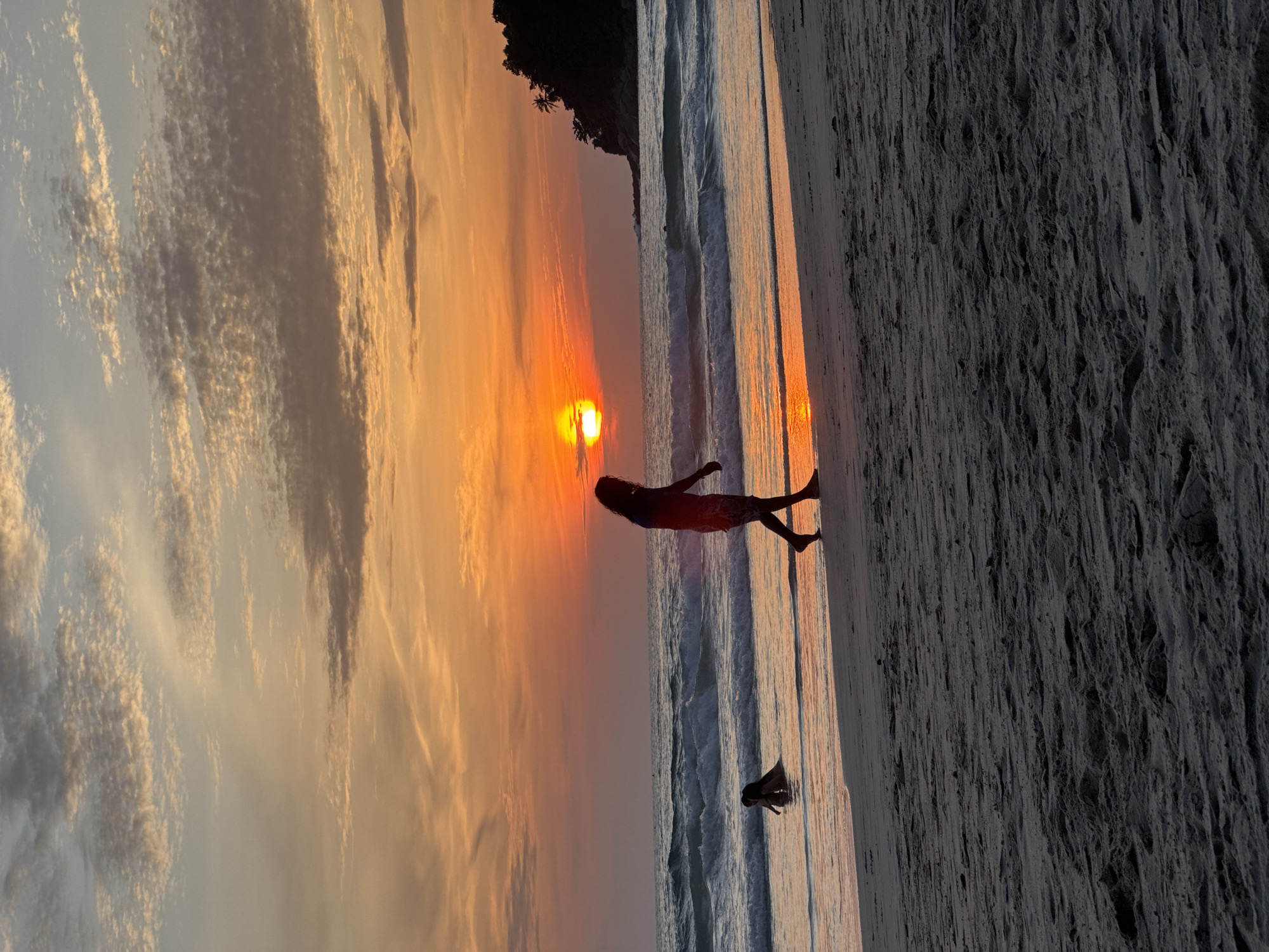 A silhouetted person walks along the sandy shore of Karon Beach, Thailand, during a vibrant sunset. The sky displays a gradient of orange and yellow hues, with scattered clouds. The ocean is visible with gentle waves breaking near the shore. In the shallow water, another silhouetted figure appears to be wading or playing. The foreground is dominated by the textured sand of the beach, with the darker tones suggesting the fading light. A distant headland with trees is visible on the right side of the image, silhouetted against the colorful sky.